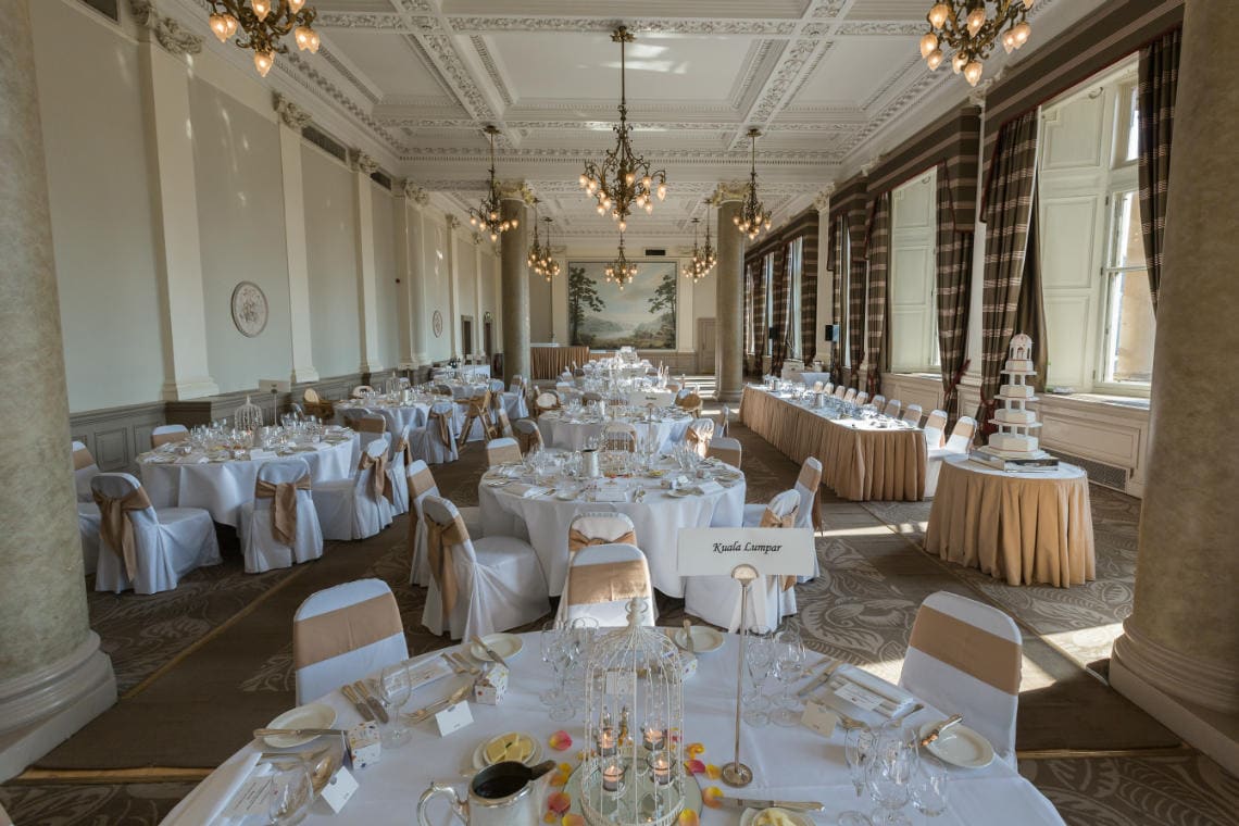 Balmoral Hotel wedding photographer image of Sir Walter Scott Suite: A grand, elegantly decorated banquet hall with round tables covered in white cloths and beige chair covers. Chandeliers hang from the ceiling, and a tiered cake sits on a table in the corner.