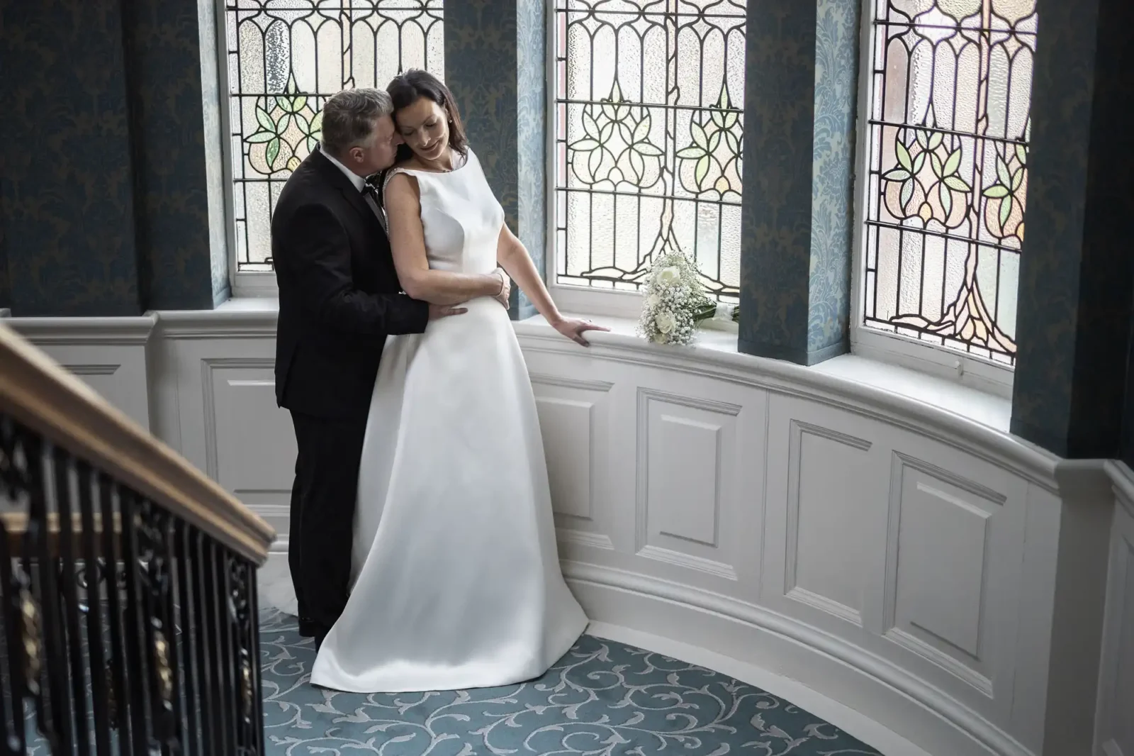 A bride in a white dress and a groom in a black suit embrace on a curved staircase with ornate patterned carpet. Behind them are tall stained glass windows and white paneled walls. A small bouquet sits on the ledge next to them.