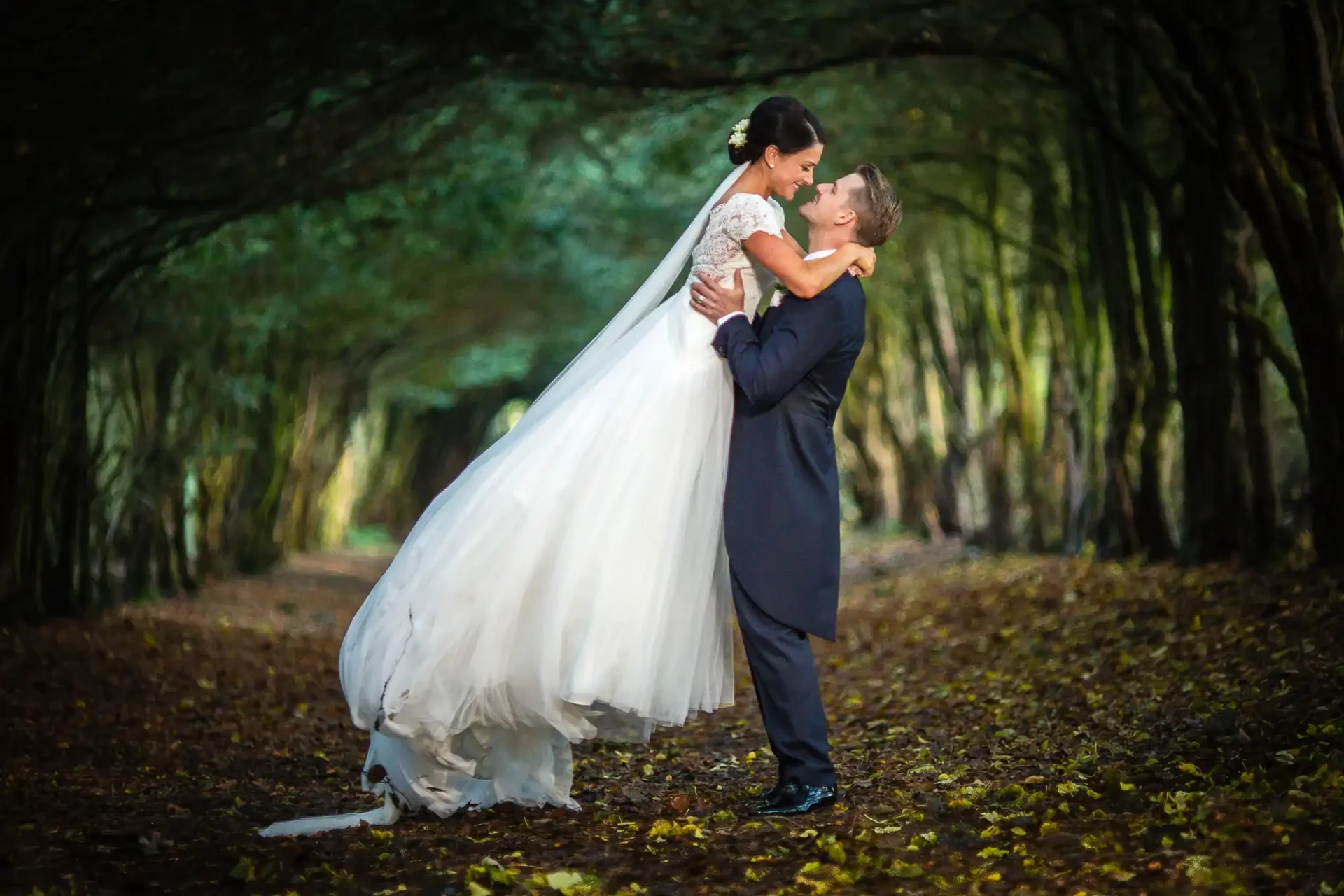 Groom lifting bride in a forested path at Barony Castle, kissing under a canopy of trees with autumn leaves on the ground.