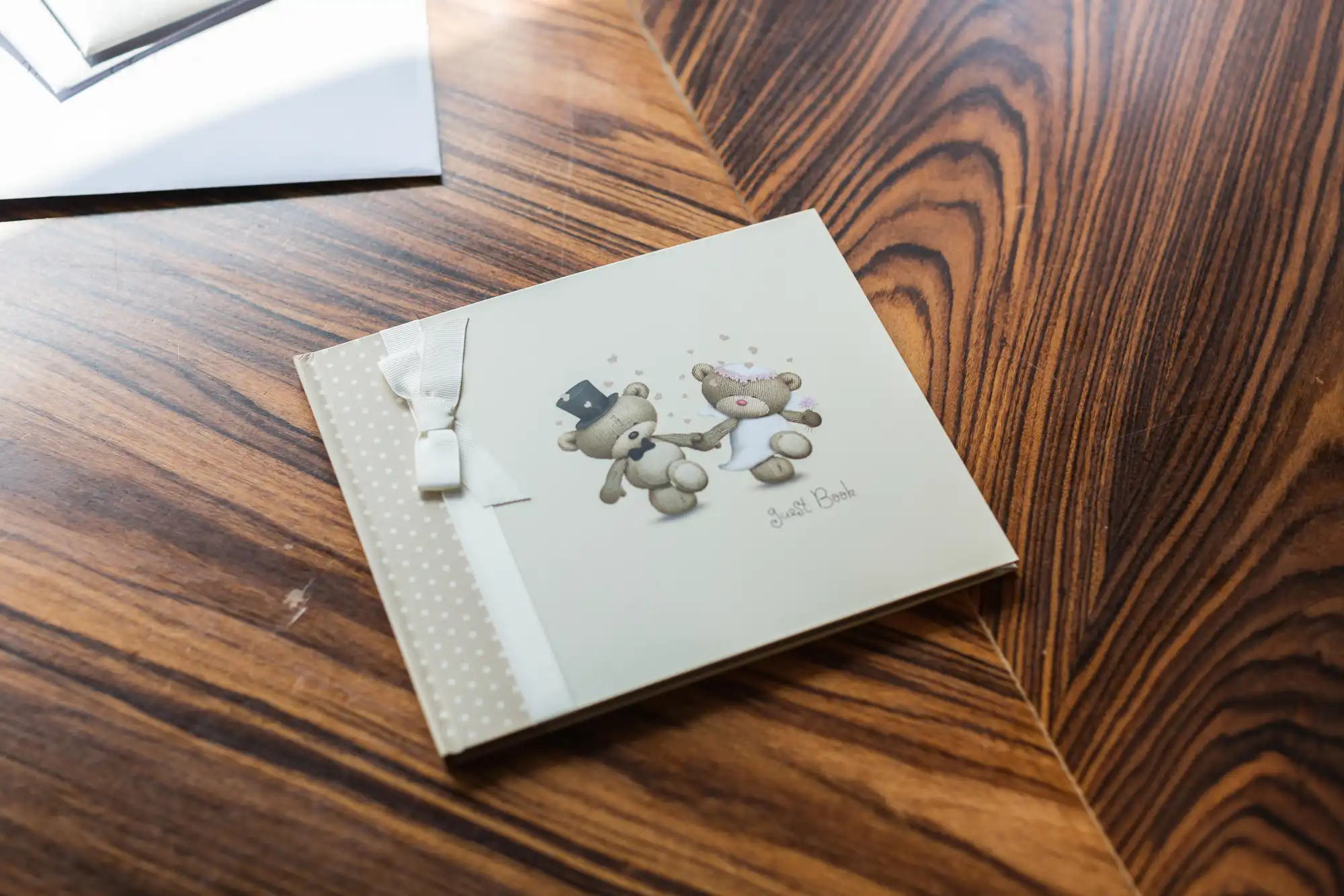 A white invitation card with a ribbon and an illustration of three teddy bears, lying on a wooden table.