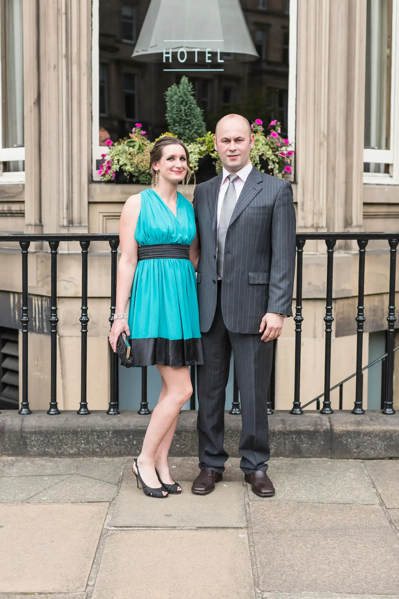 A couple standing in front of a hotel entrance, with the woman dressed in a teal dress and the man in a gray suit.