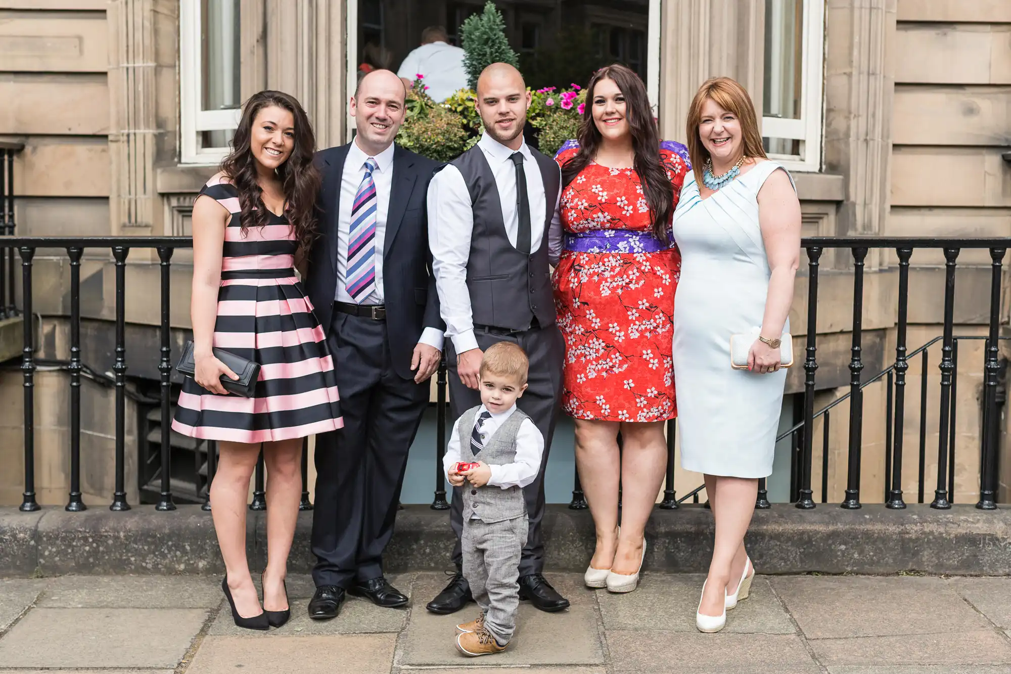 A family of six, including a toddler, dressed in formal attire, posing together in front of a building.