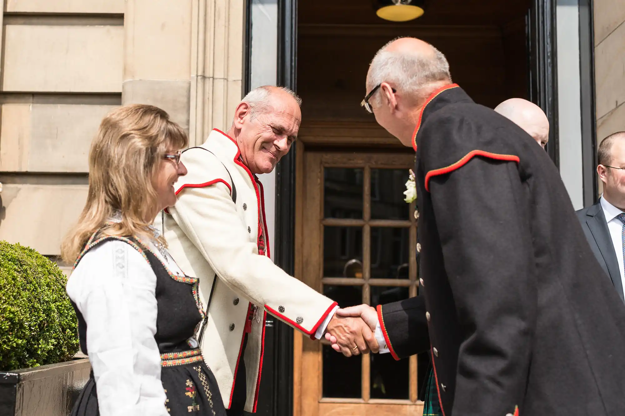 Two men in traditional attire shaking hands and smiling at a formal event, with a woman looking on.