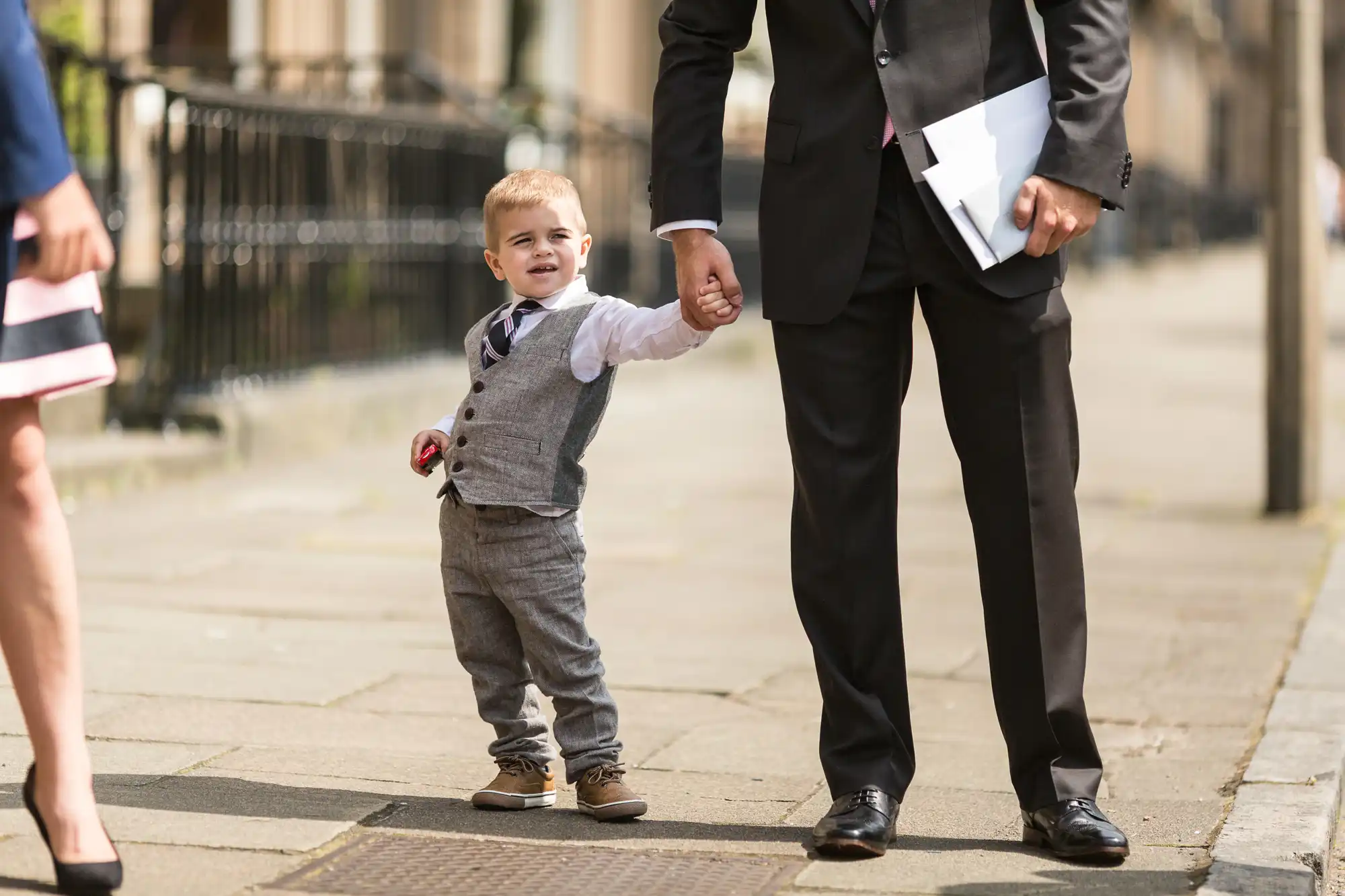 A toddler in a gray vest and pants holding hands with an adult in a suit on a sunny city street, with woman's legs visible on the left.