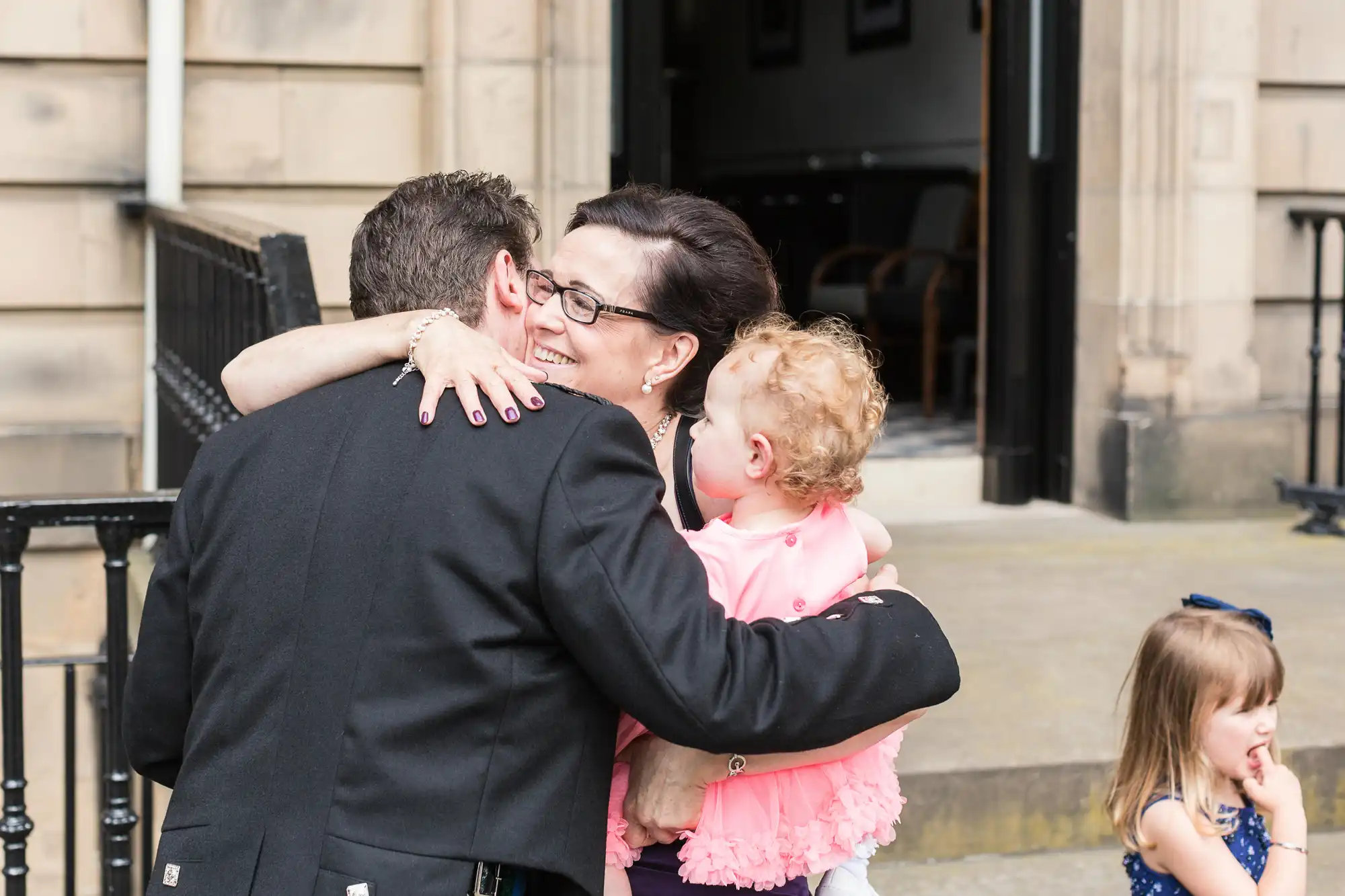 A woman hugs a man, holding a toddler in a pink outfit, while another child stands nearby, on a city sidewalk.