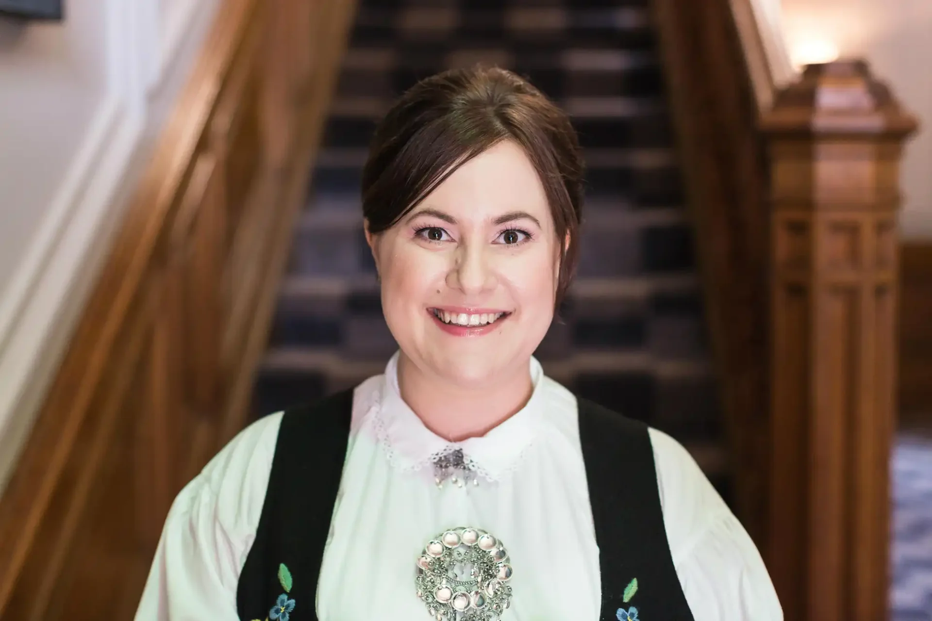 A woman in traditional folk attire smiling at the camera, standing in front of a staircase.