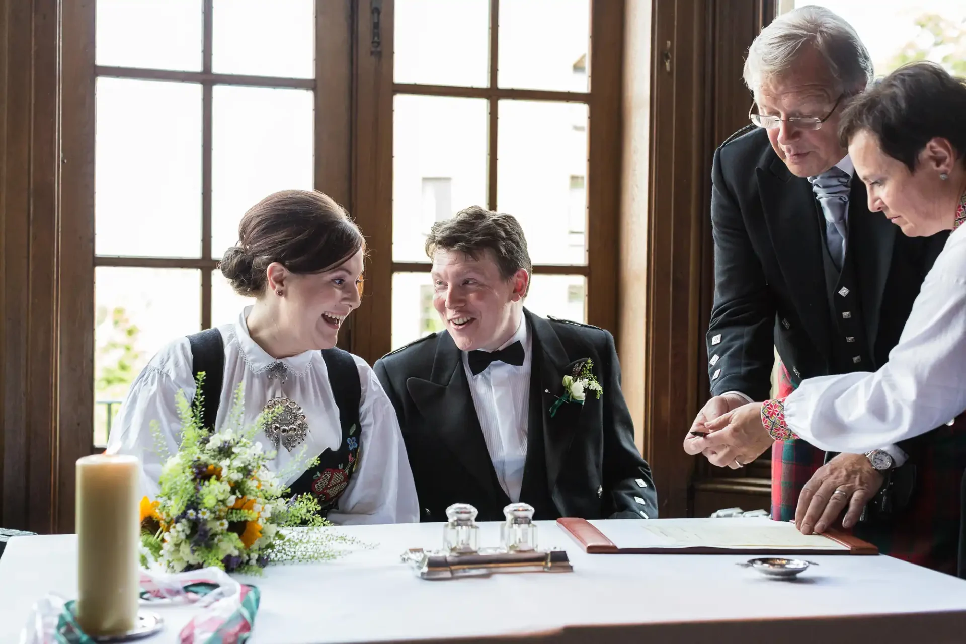 A newlywed couple in traditional attire signing their marriage certificate, surrounded by older family members, at a table with a floral centerpiece.