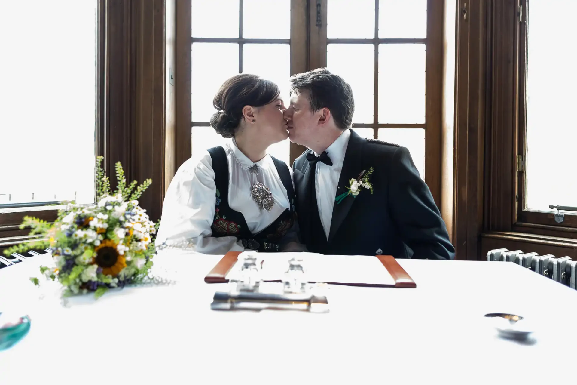 A bride and groom in traditional attire kiss at a table in a well-lit room, with a bouquet and signing documents in front of them.