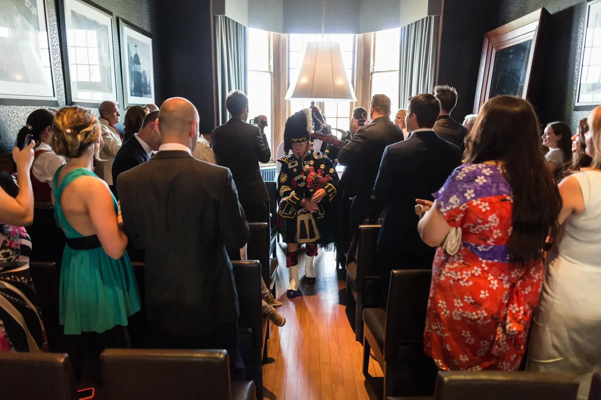 A bagpiper in traditional scottish attire leads a procession through a crowd of formally dressed guests at an indoor event.