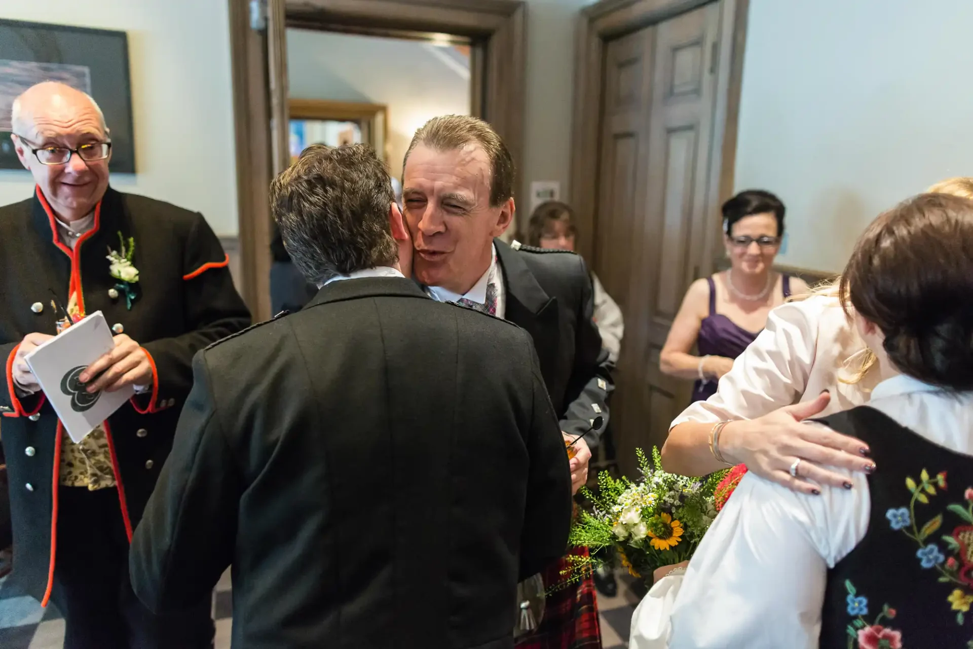 Two men in suits embrace warmly at an indoor gathering, while a woman in floral dress and others watch.