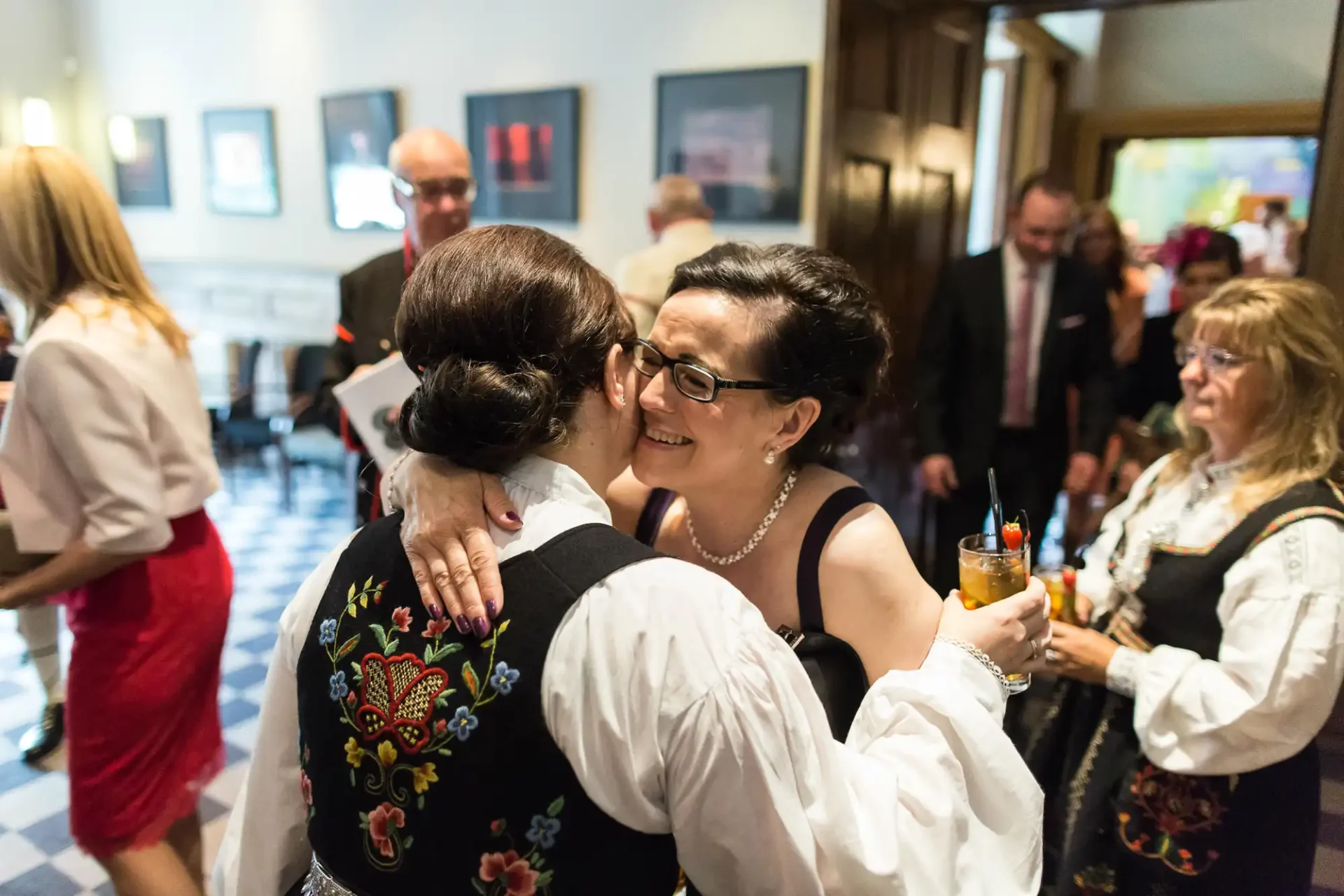 Two women embracing warmly at a social gathering, one holding a drink, surrounded by other guests in a cheerful atmosphere.