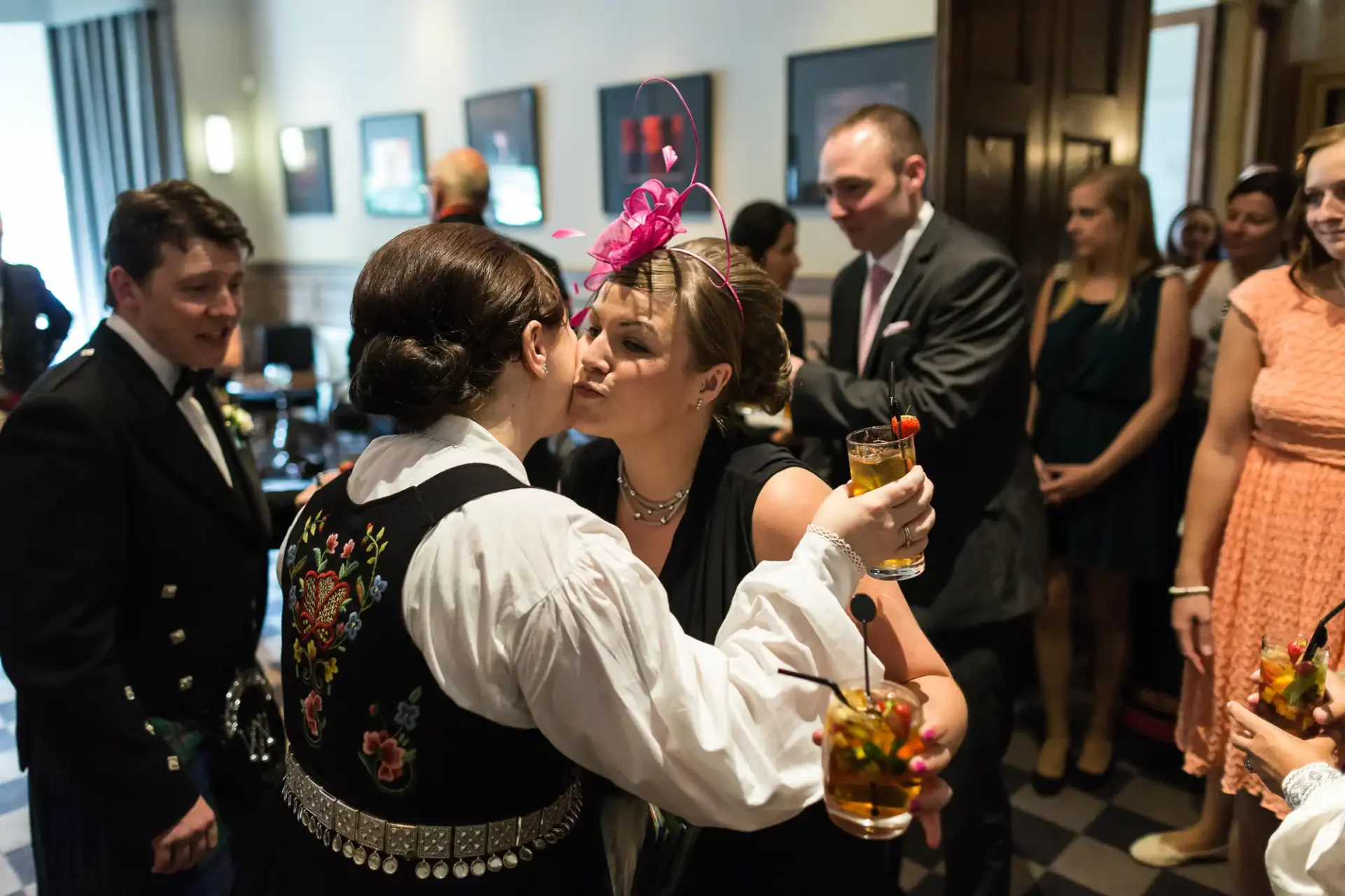 Two women in a festive gathering, one in traditional embroidered dress, kiss on the cheek while holding drinks, surrounded by onlookers.