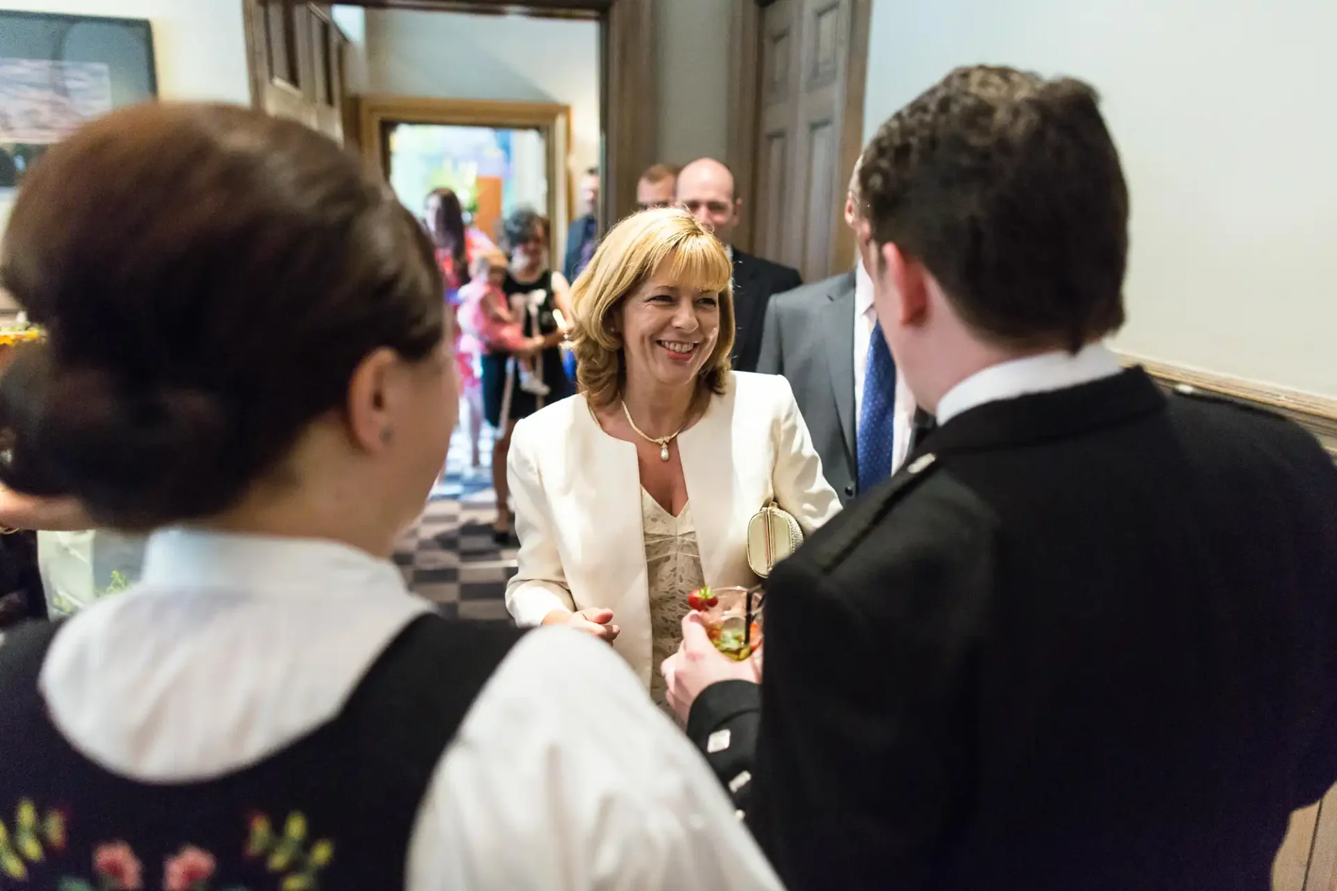A woman in a white suit smiling at two servers during an indoor event.