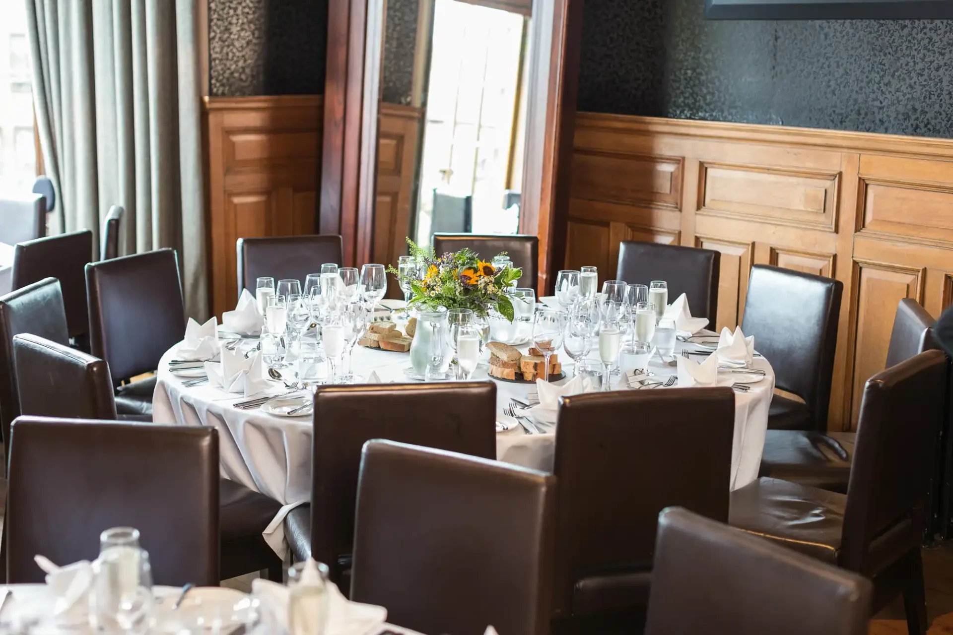 Elegant dining setup with a round table, white linen, a floral centerpiece, wine glasses, and black chairs in a room with wood paneling.