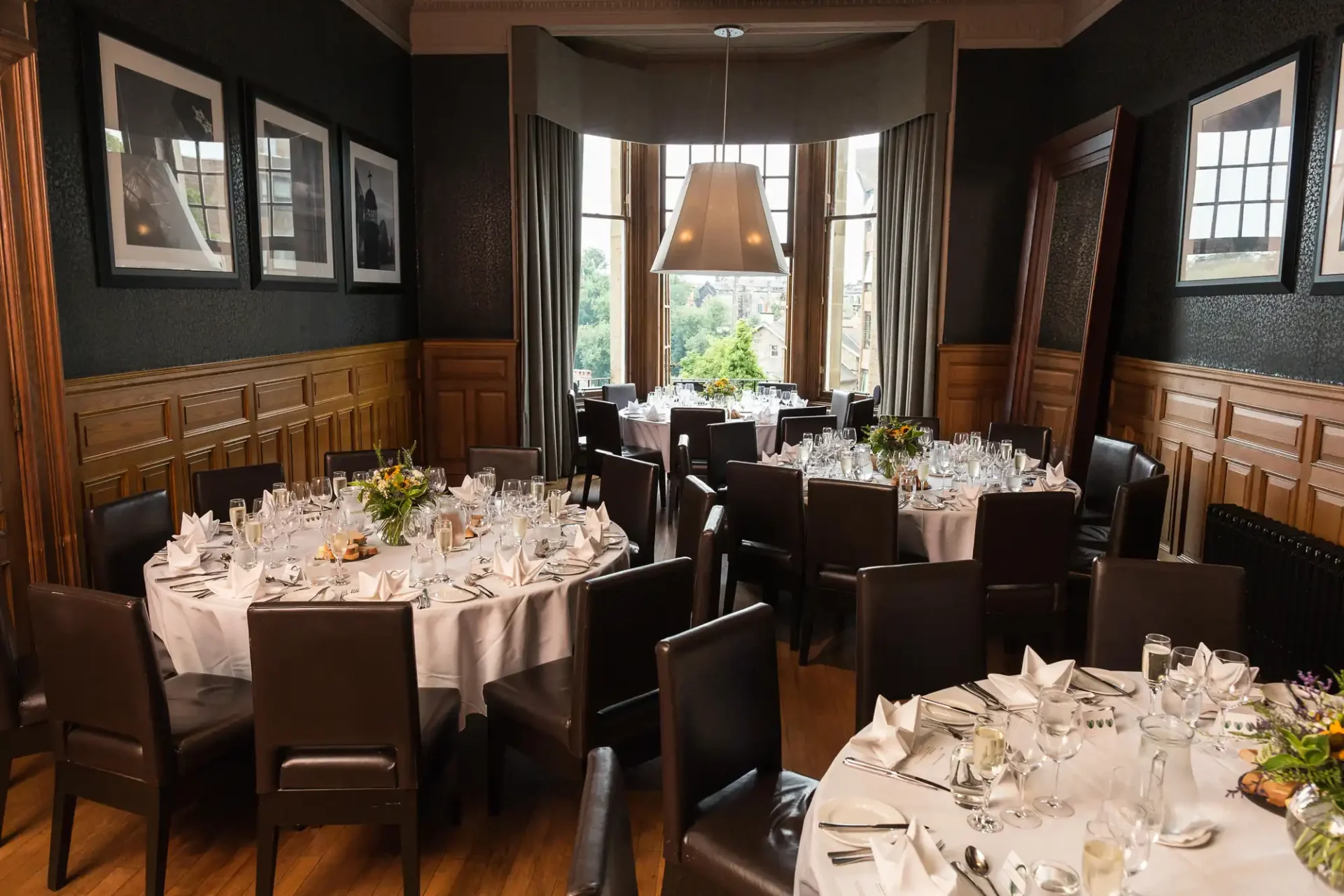 Elegant dining room with round tables set for a formal meal, featuring white tablecloths, floral centerpieces, and framed photographs on dark walls.