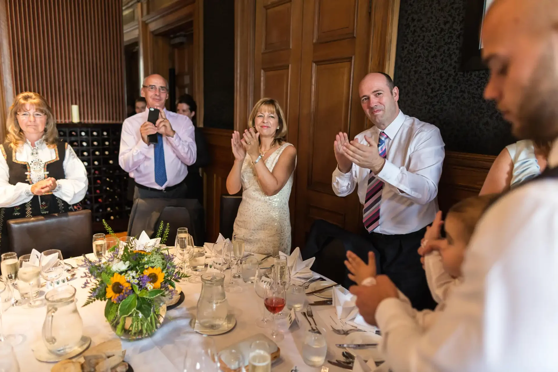 Guests at a wedding reception clapping, seated around a decorated table with a floral centerpiece in an elegantly paneled room.