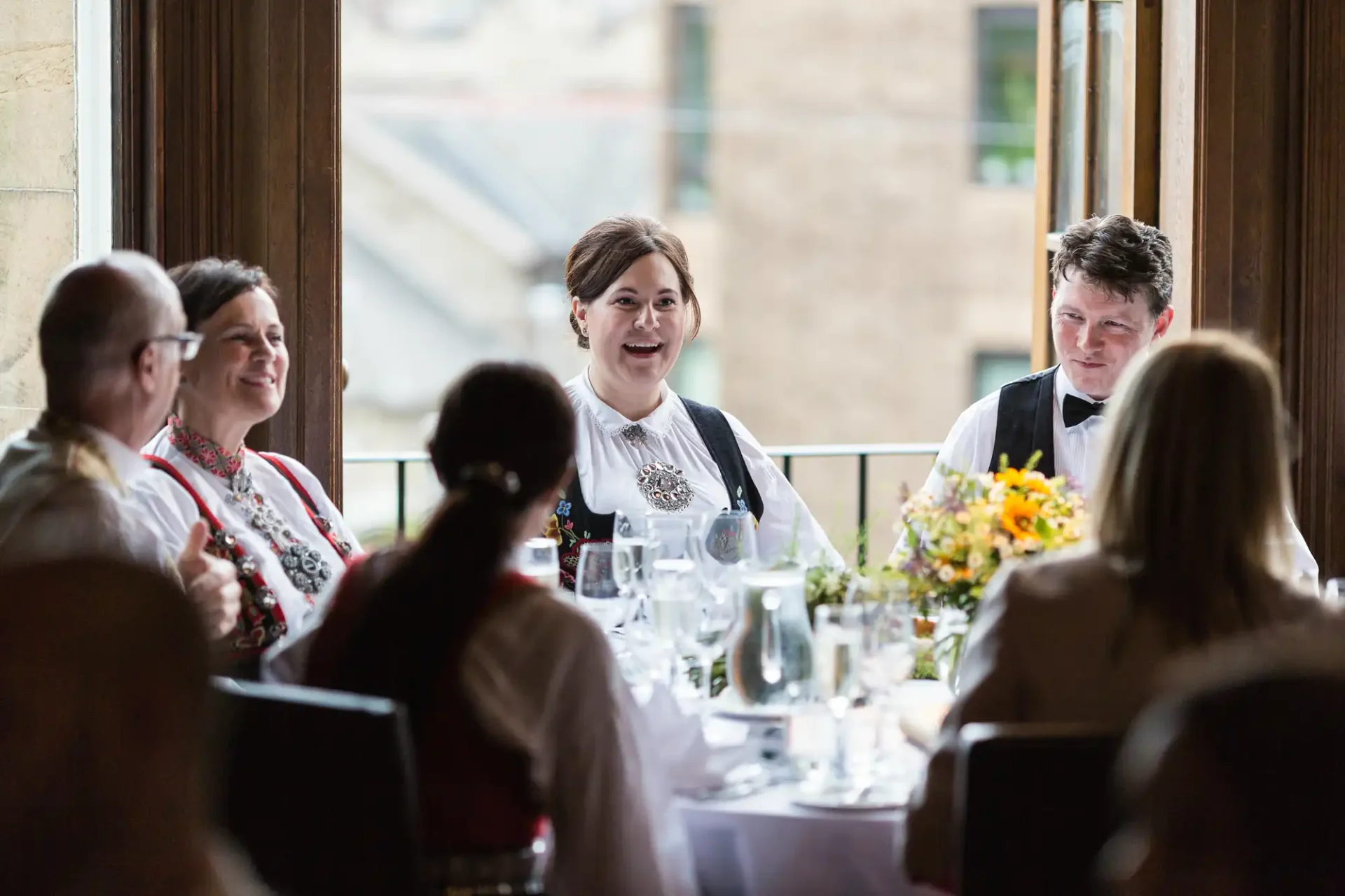 A group of people enjoying a lively conversation around a dinner table with elegant table settings and a flower centerpiece.