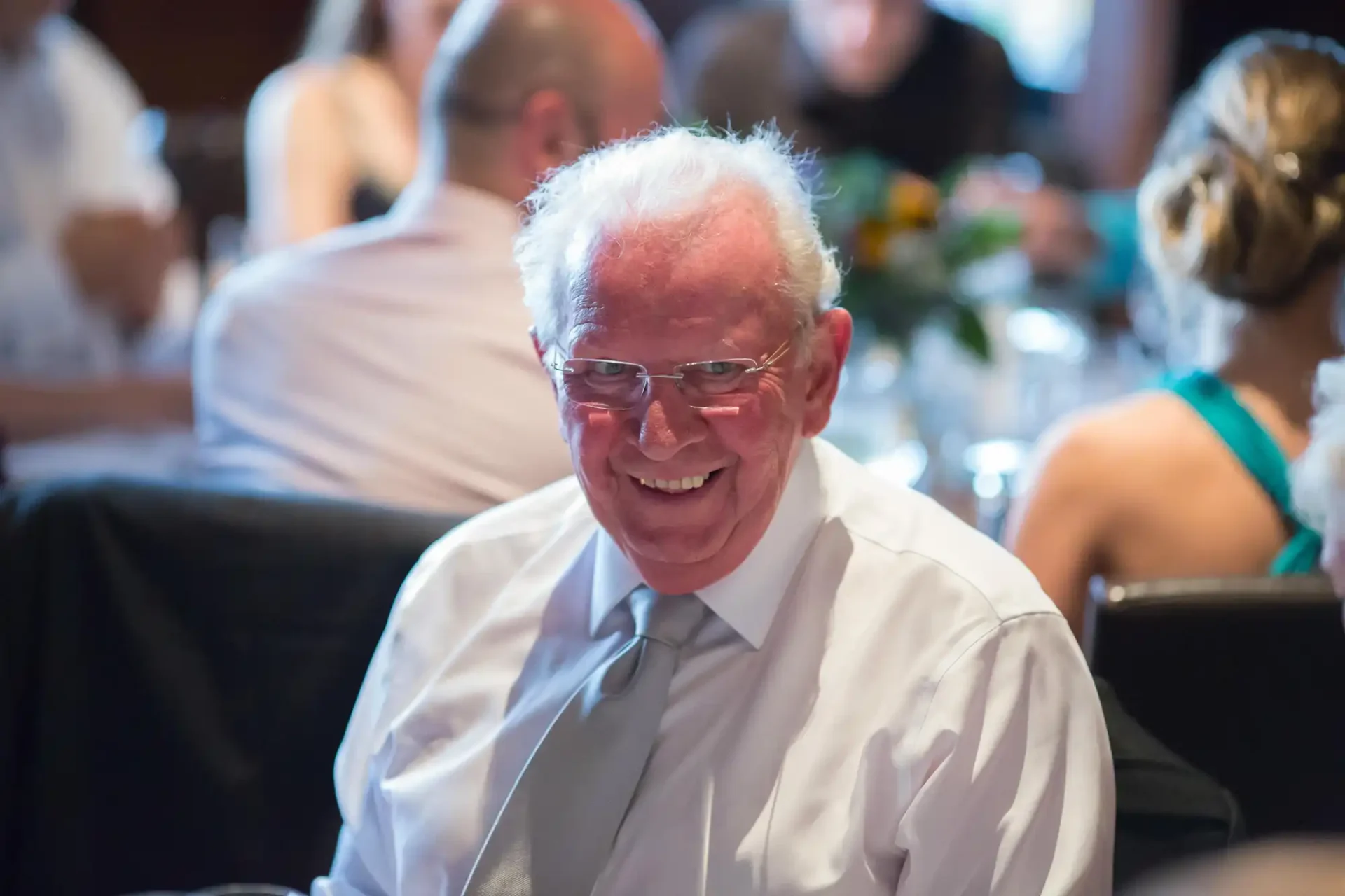 Elderly man with white hair smiling at a formal gathering, wearing a white shirt; blurred people in the background.