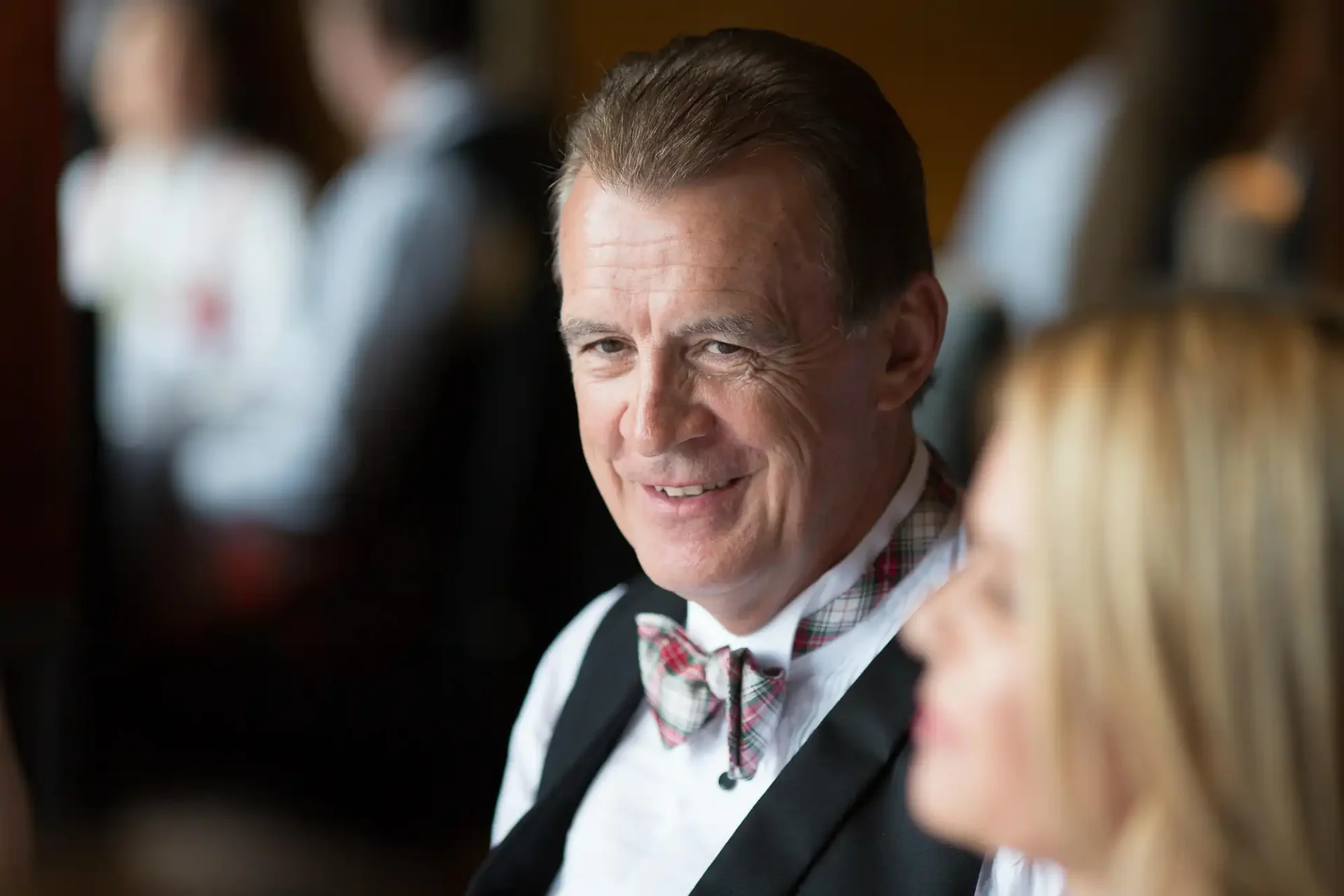 A smiling middle-aged man in a tuxedo and bow tie, with a woman partially visible in the foreground, both indoors.