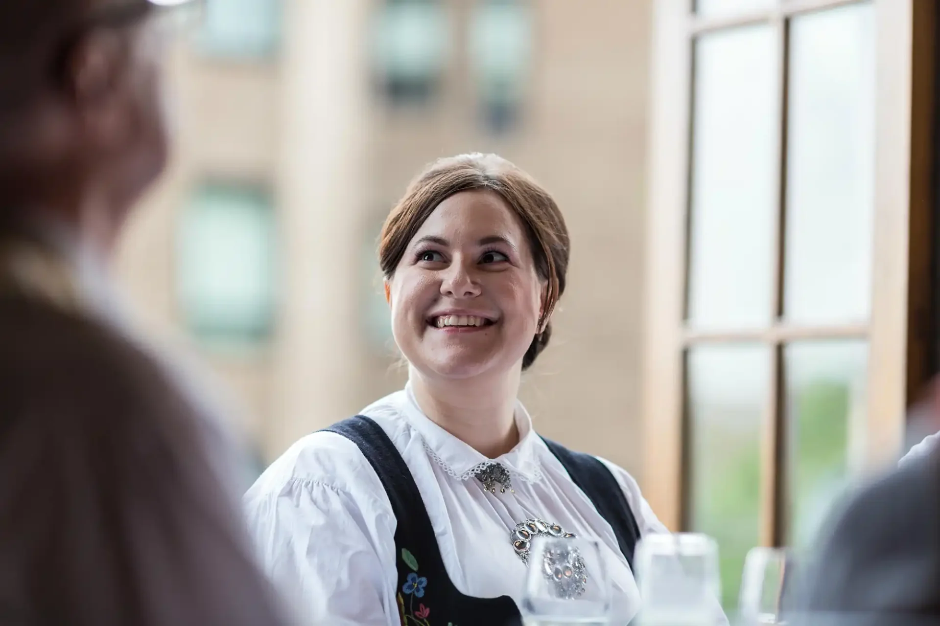 A woman in traditional attire smiling during a conversation at a social gathering, with a blurred background of windows and another person.