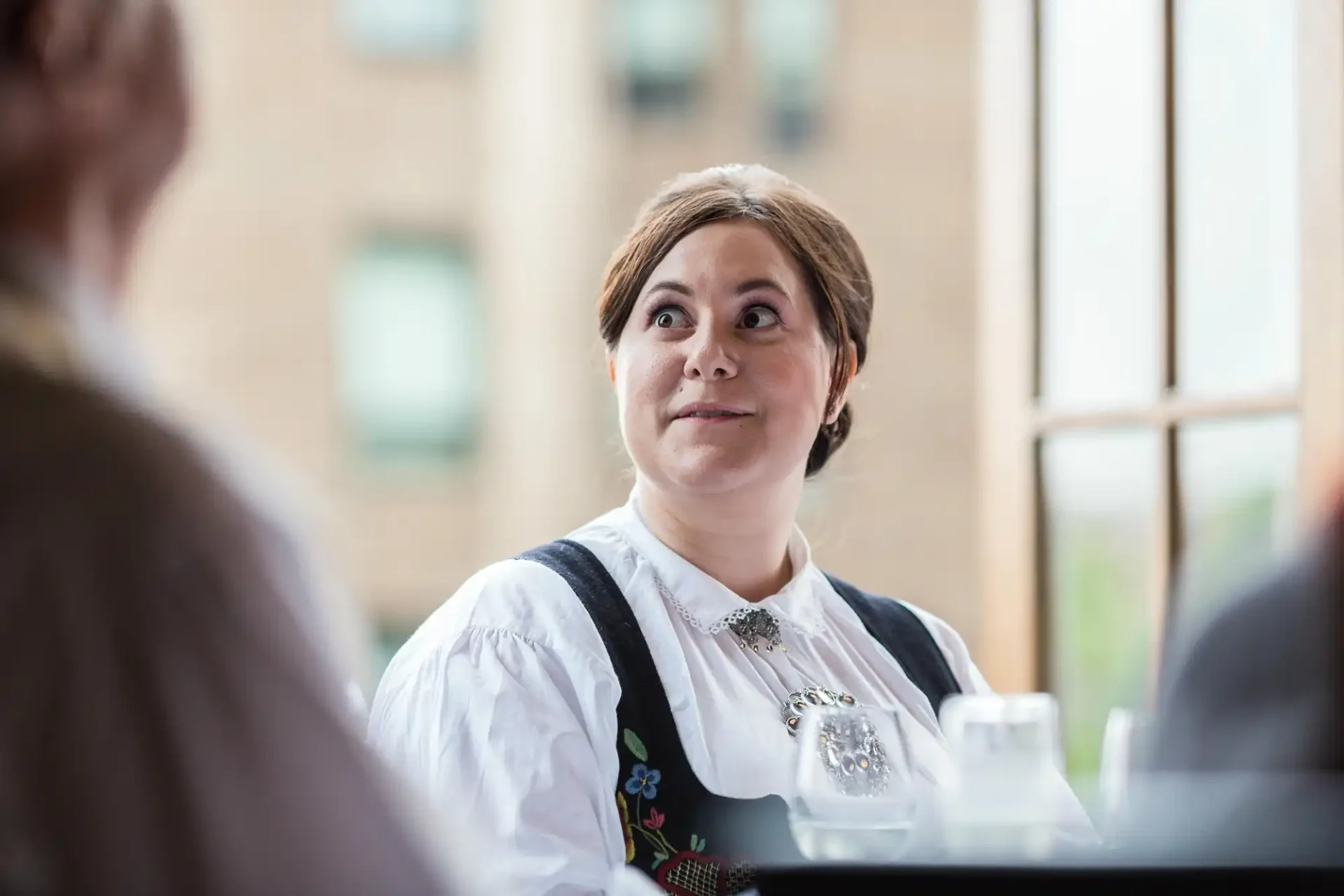A woman in traditional dress makes an amusing facial expression while conversing with someone out of frame in a brightly lit space.