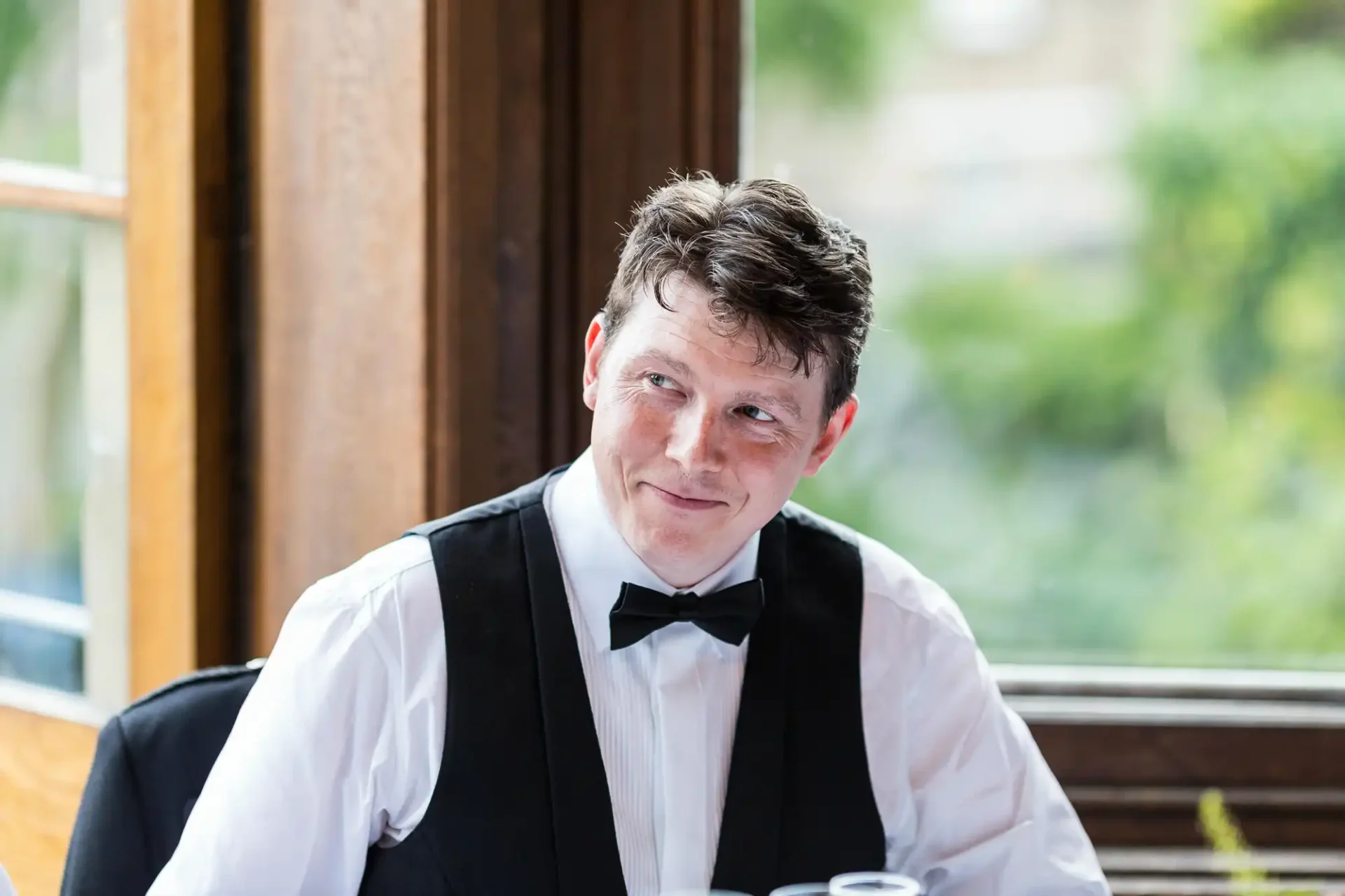 A man in a black vest and bow tie smiles while sitting indoors near a window, with a blurred green background visible outside.