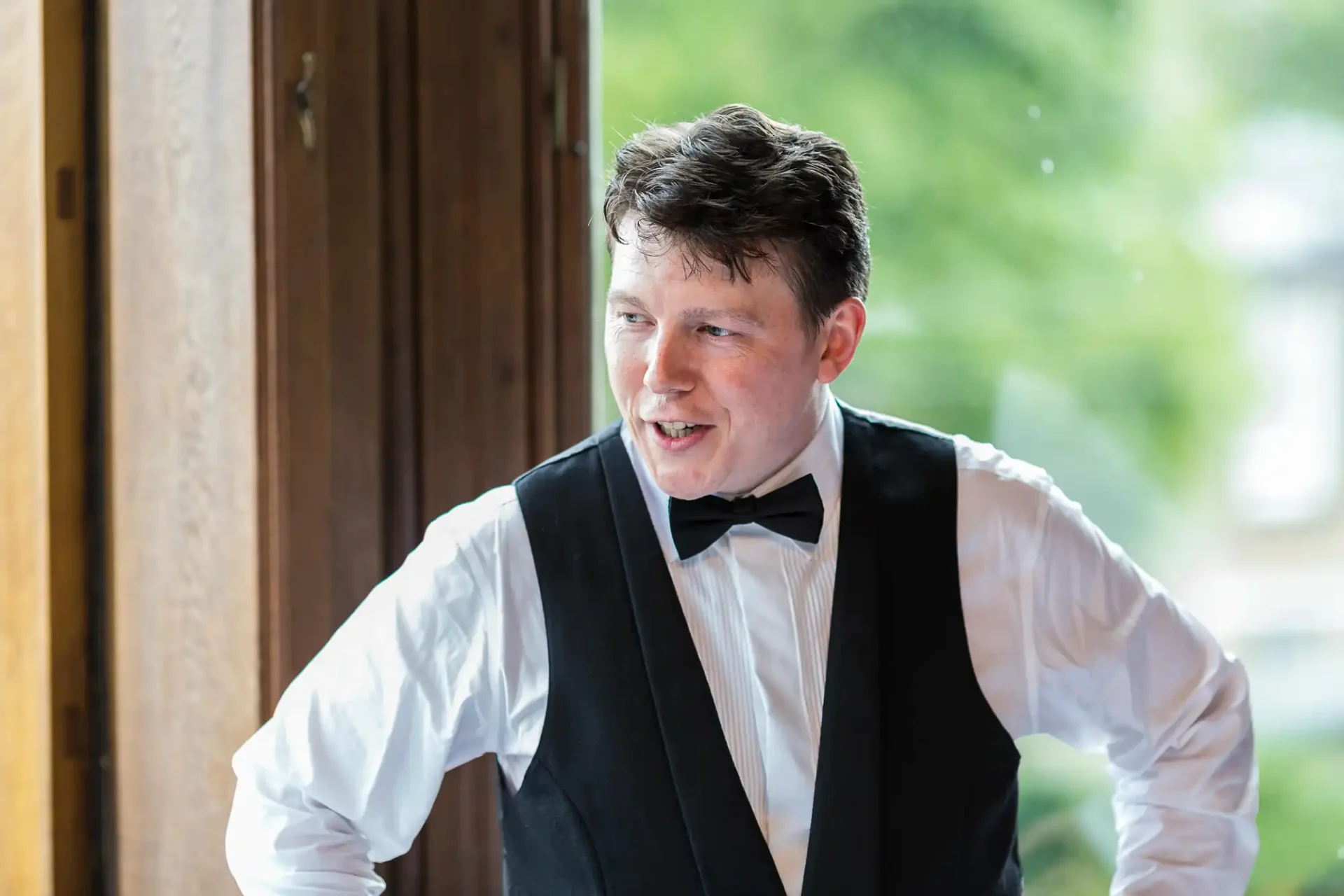 Man in a tuxedo smiling and leaning forward, standing by a wooden door with blurred bright background.