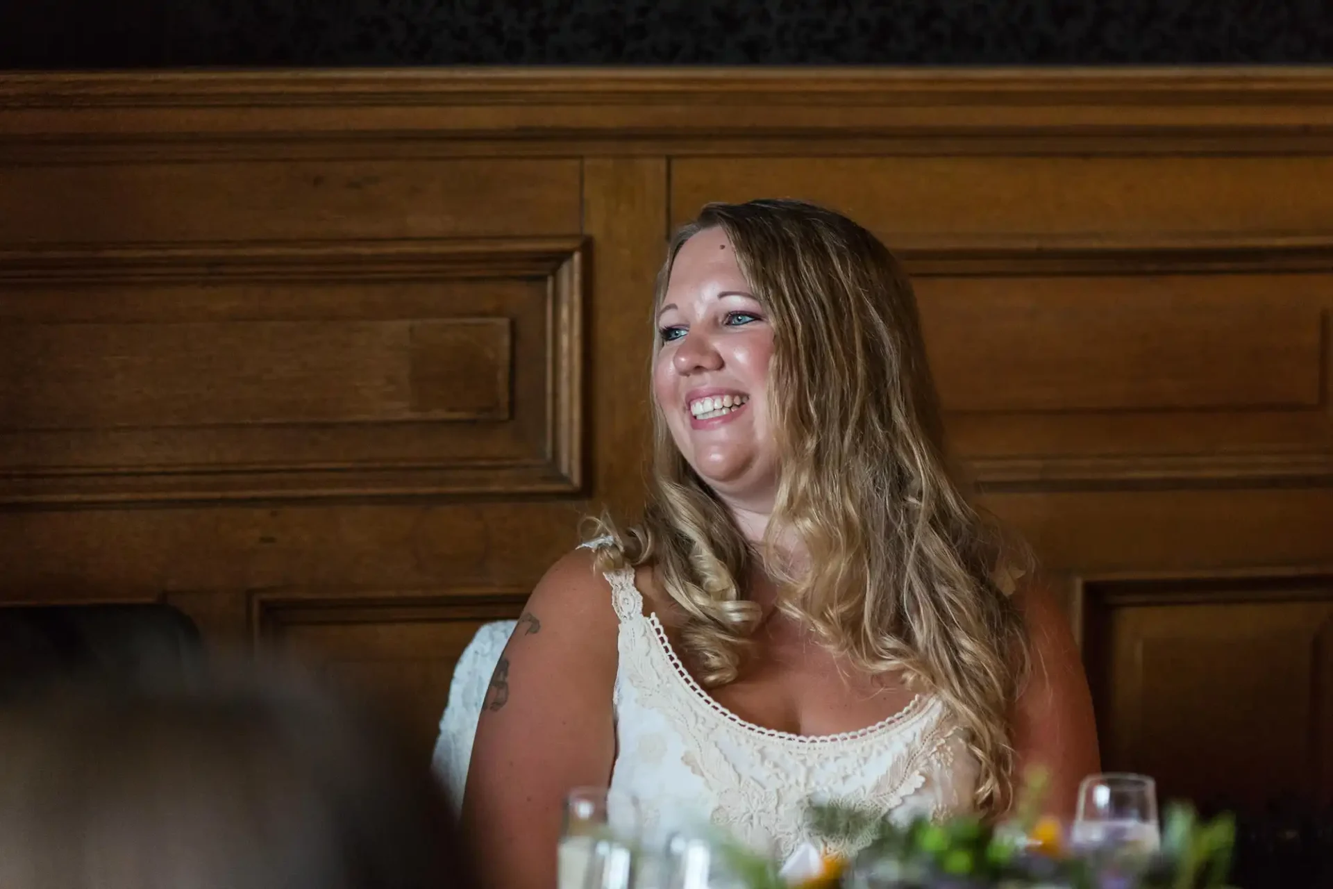 A smiling woman with long blonde hair, wearing a white dress, sitting at a dark-wood table in a dimly lit room.