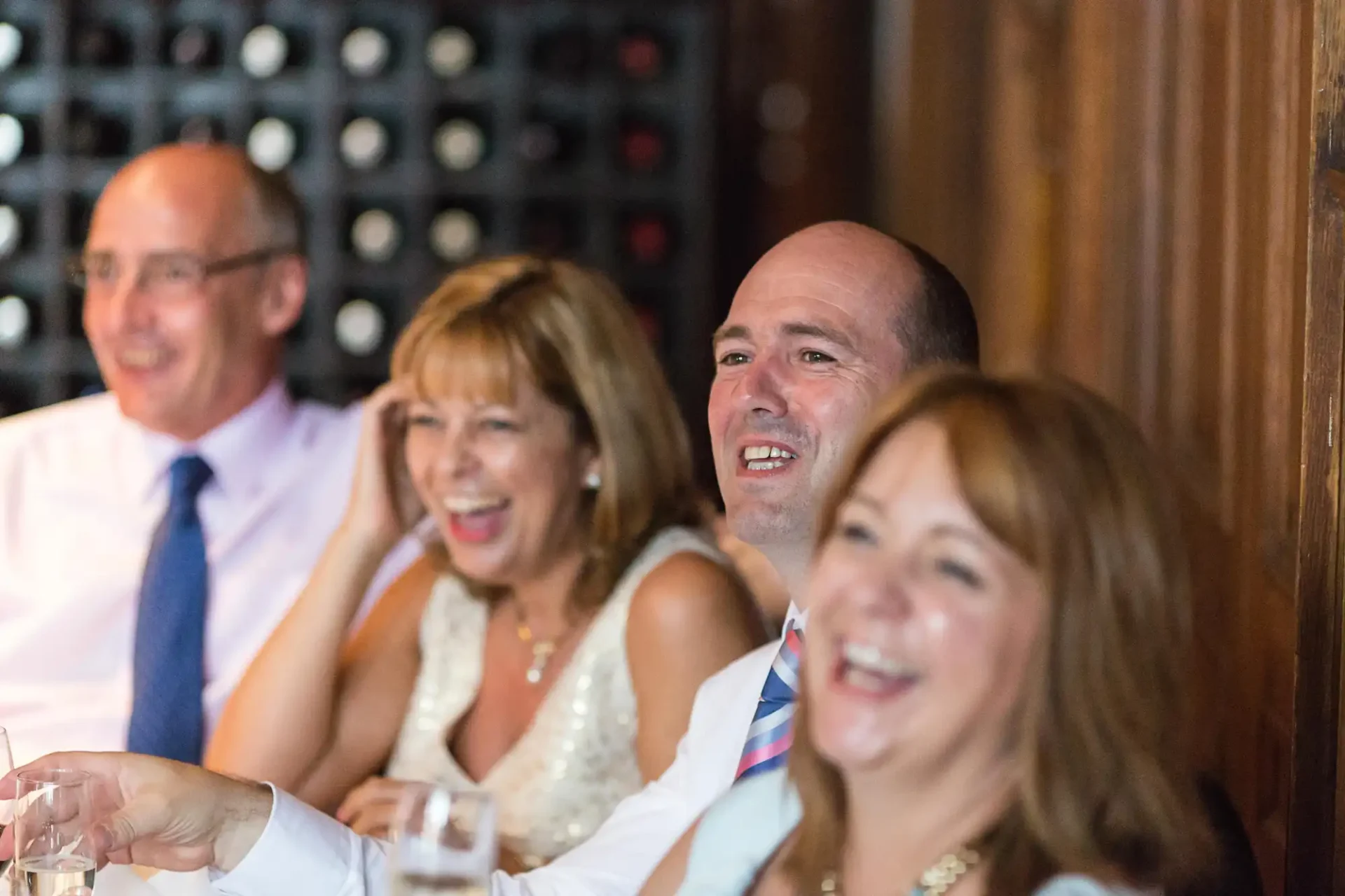 Four adults laughing and enjoying themselves at a social gathering, seated at a table with wine bottles in the background.