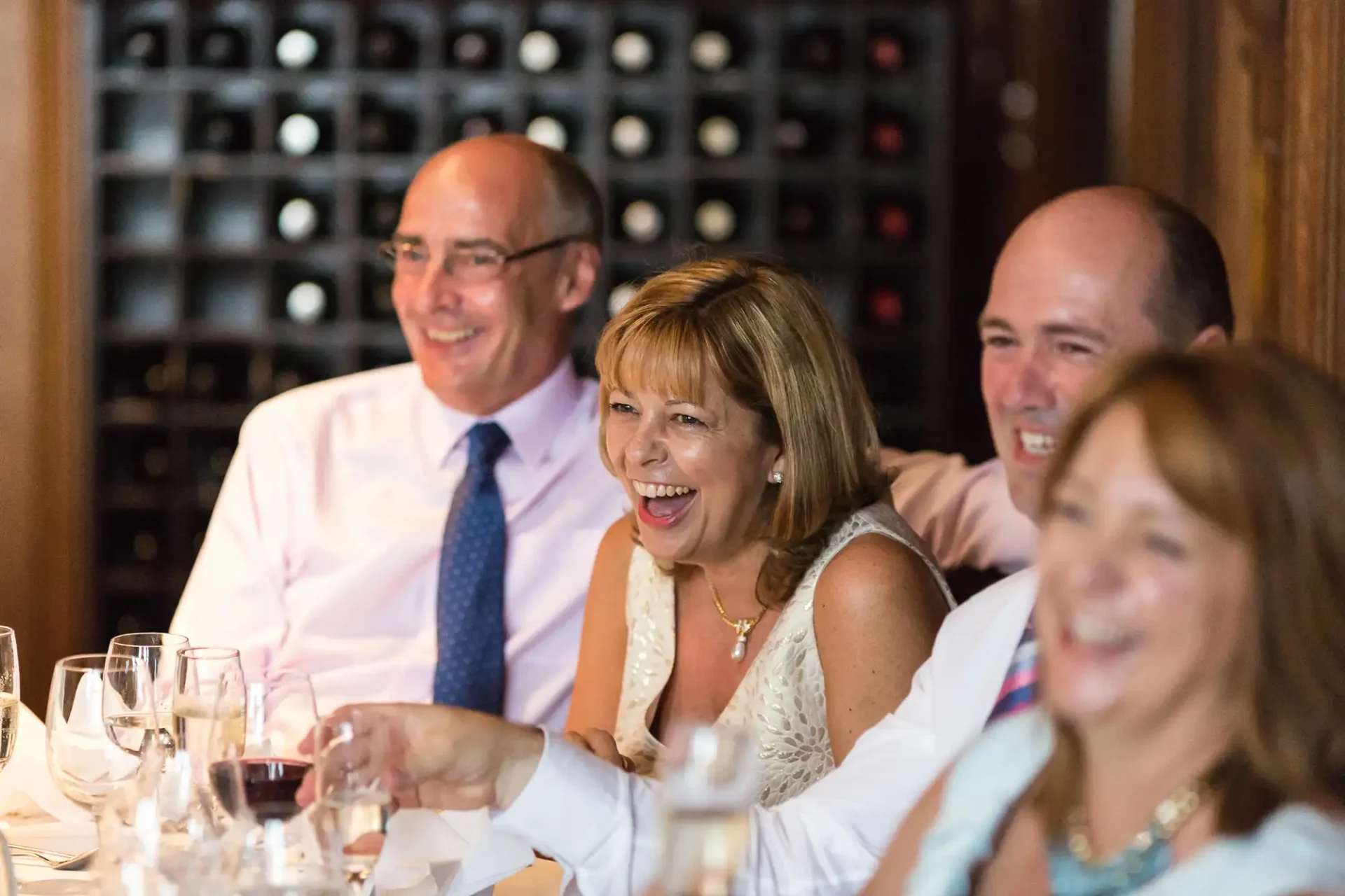 Two men and two women laughing and enjoying a meal together at a table in a restaurant with wine bottles in the background.