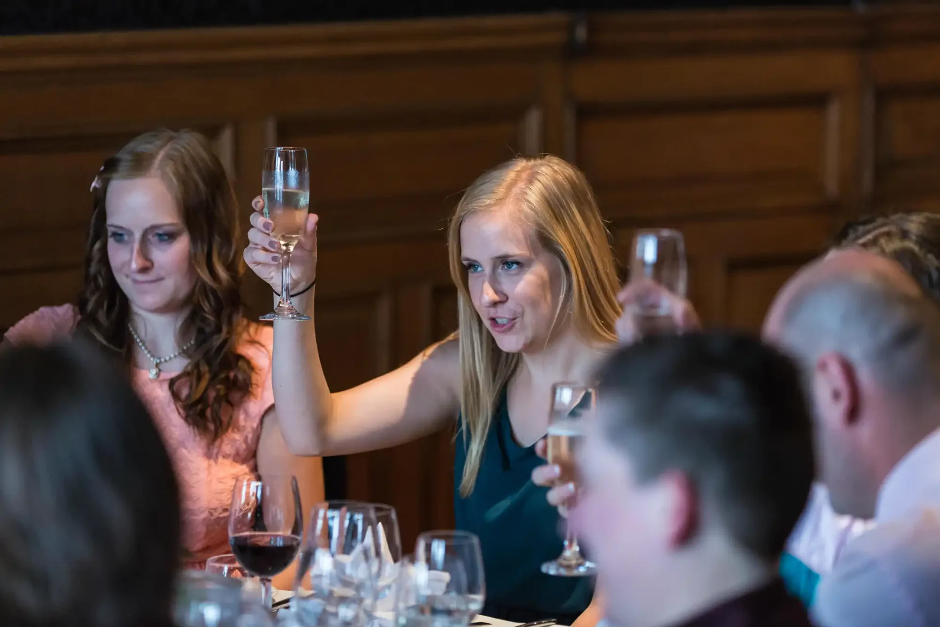 Woman raising a toast with a champagne glass at a dining table surrounded by guests in a warmly lit room.