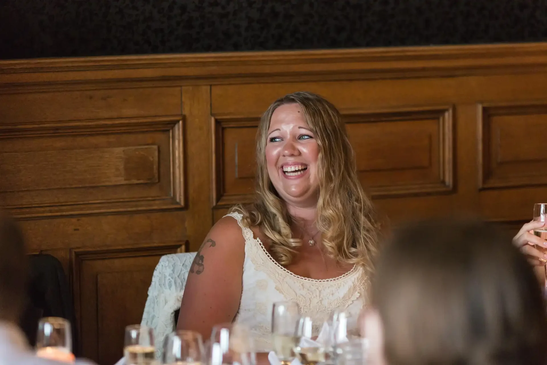 A joyful woman laughing at a dining table during a formal event, with a glass in her hand and elegant wooden paneling in the background.