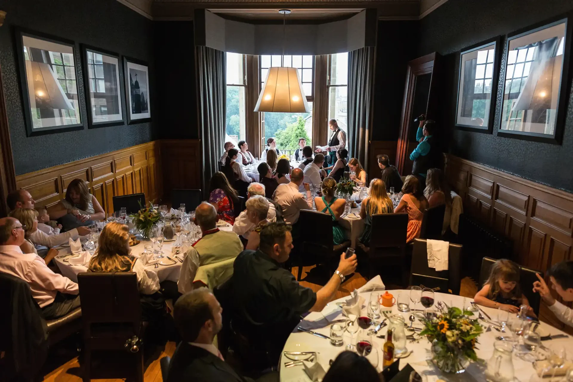 A wedding reception in a dimly lit room with guests seated around tables, listening to a speech by a standing couple.