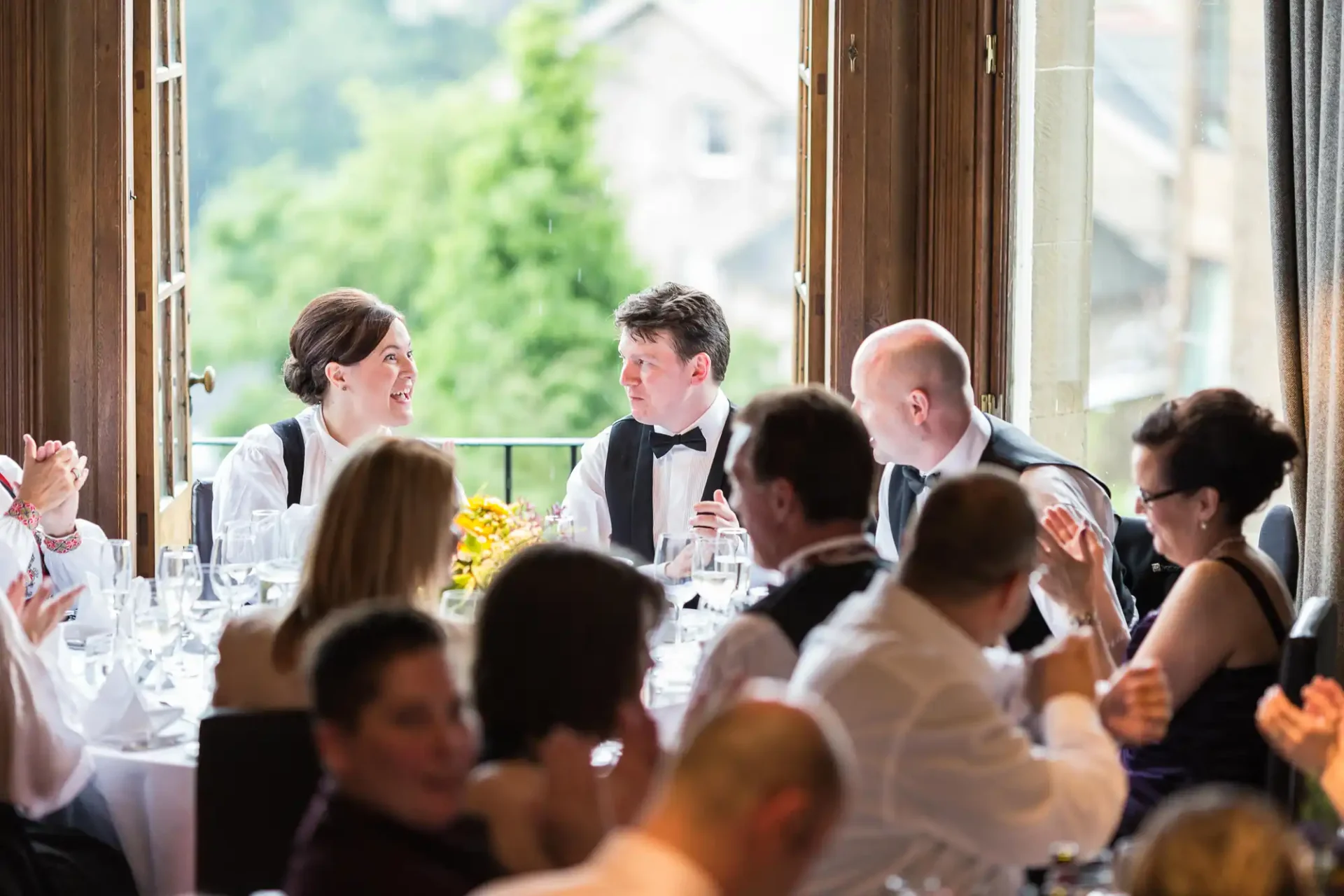 A couple in formal wear engaging in conversation at a wedding reception table surrounded by guests.