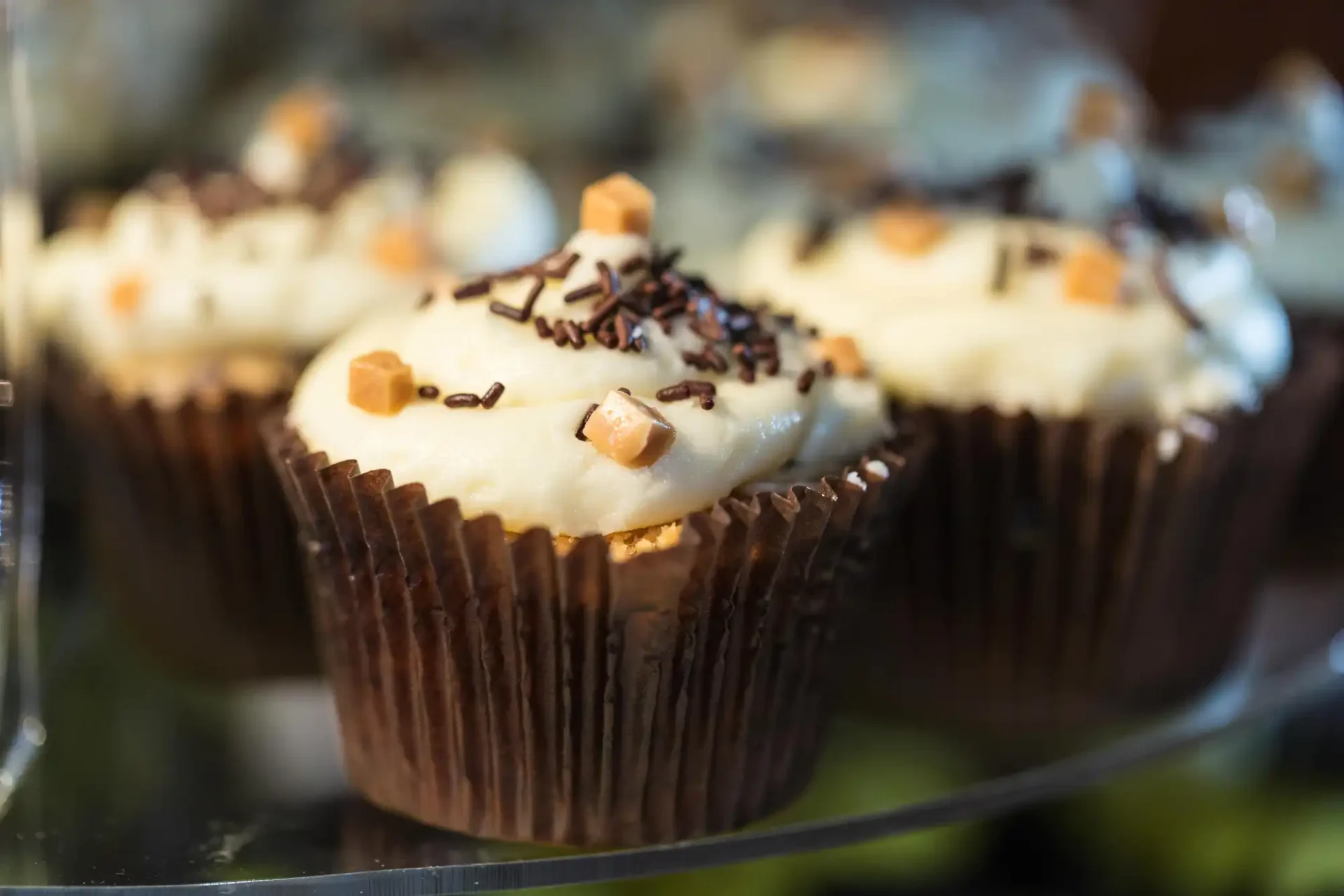Close-up of frosted cupcakes with sprinkles and diced caramel, displayed in a bakery case.