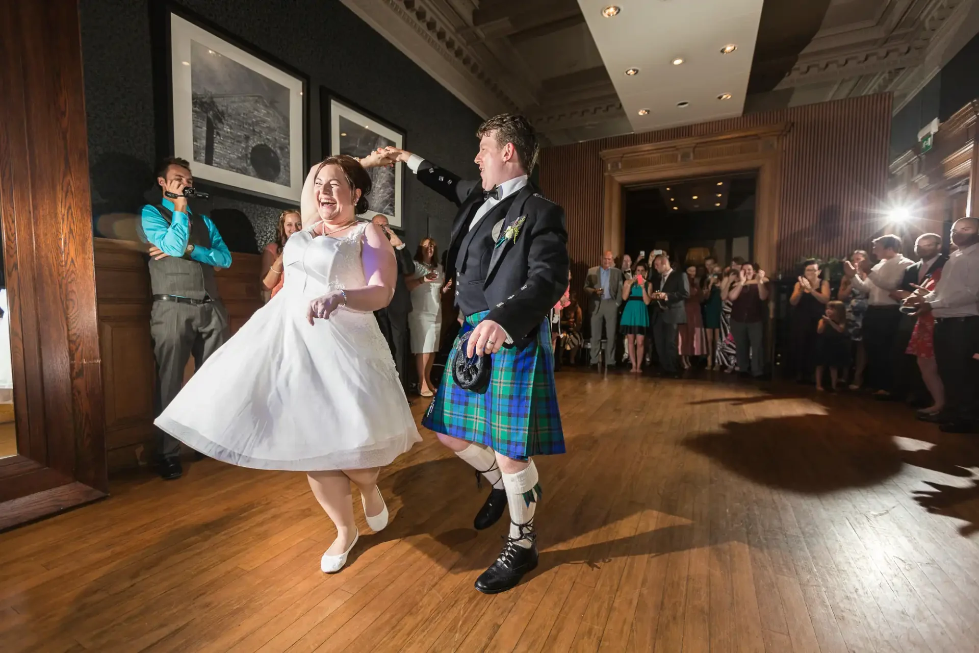 Bride and groom in traditional attire joyfully dancing at their wedding reception, surrounded by applauding guests.
