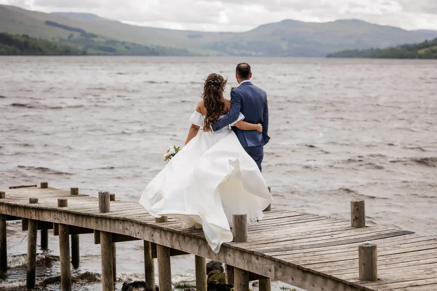 A bride and groom walk arm in arm on a wooden dock by a lake, facing the water with mountains in the background during their wedding photoshoot at Boreland Loch Tay, Fearnan, Aberfeldy.