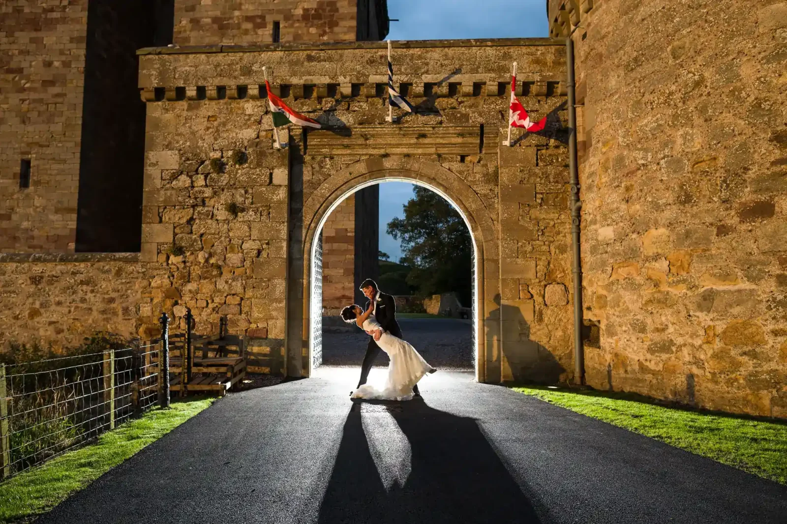 A bride and groom share a dance in front of an illuminated stone archway at night, with several flags hanging above the entrance at Borthwick Castle, Midlothian.