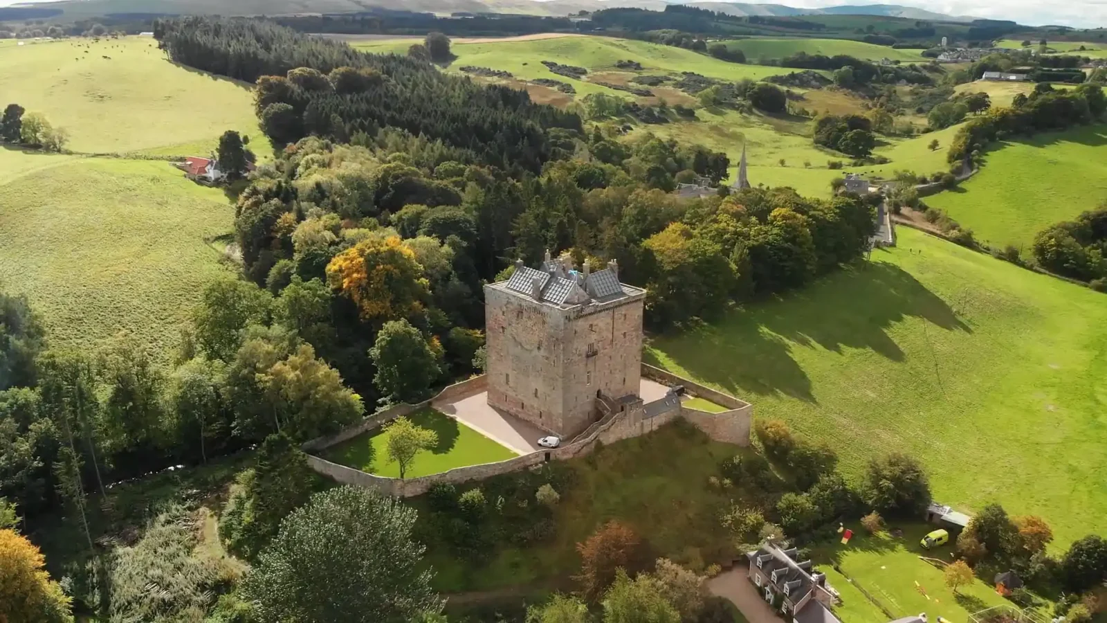 Aerial view of a stone castle surrounded by a wall, set amid green fields and trees in a rural landscape.