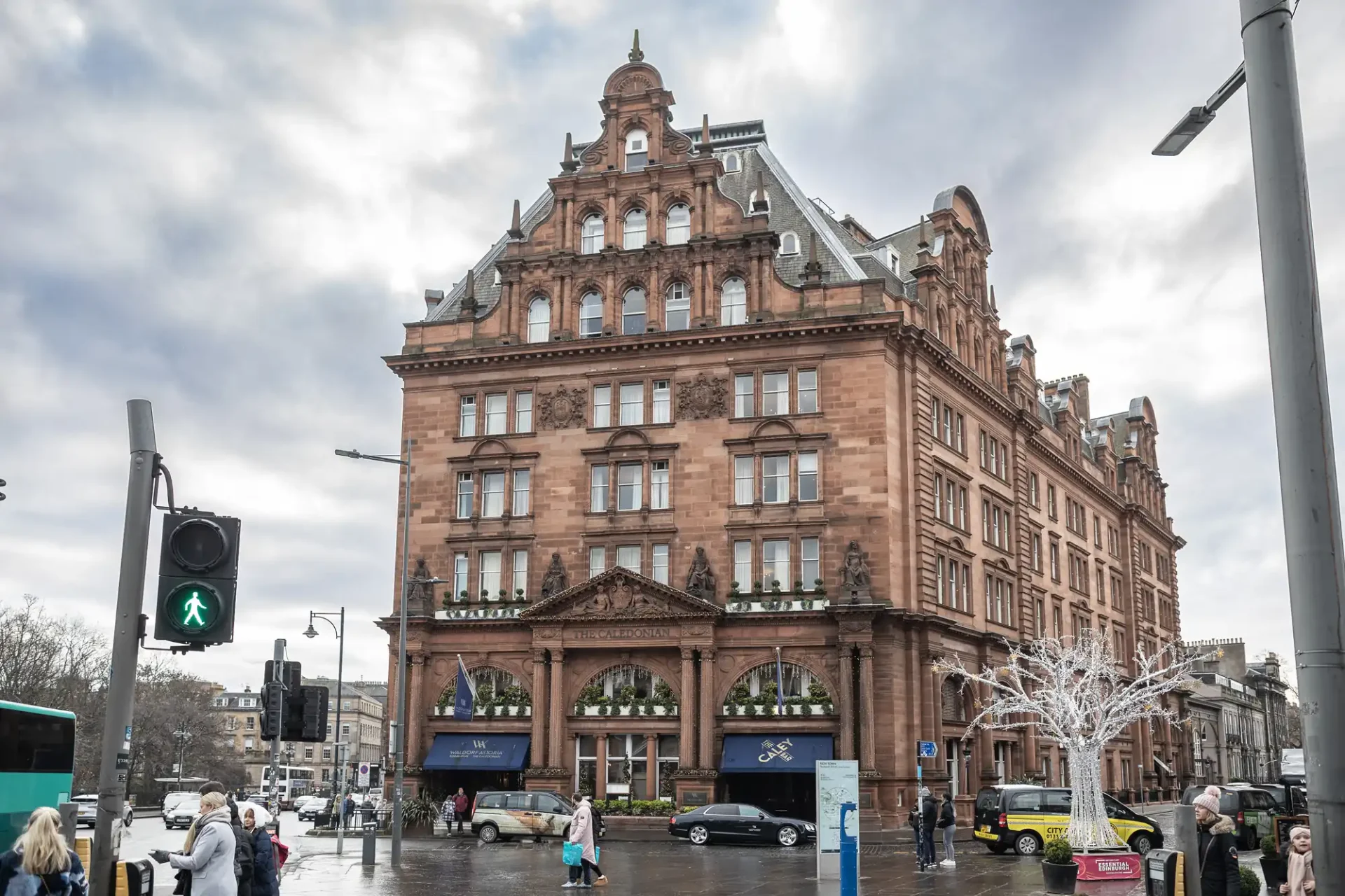 The Caledonian Edinburgh wedding photographer image of a large, historic, red-brick building on a busy city street corner, with pedestrians, a traffic light, and a few parked cars visible, under a cloudy sky.