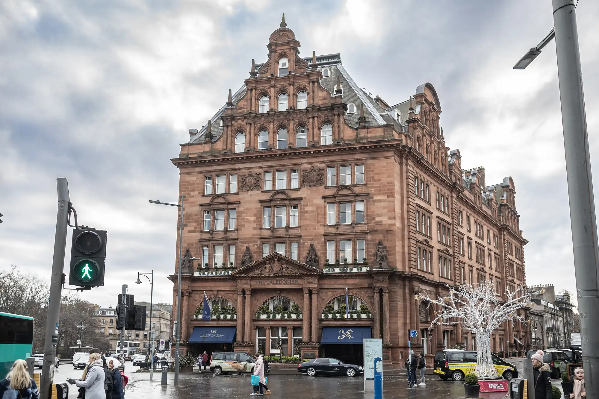 Caledonian Edinburgh wedding photographers: A large, historic, red-brick building on a busy city street corner, with pedestrians, a traffic light, and a few parked cars visible, under a cloudy sky.