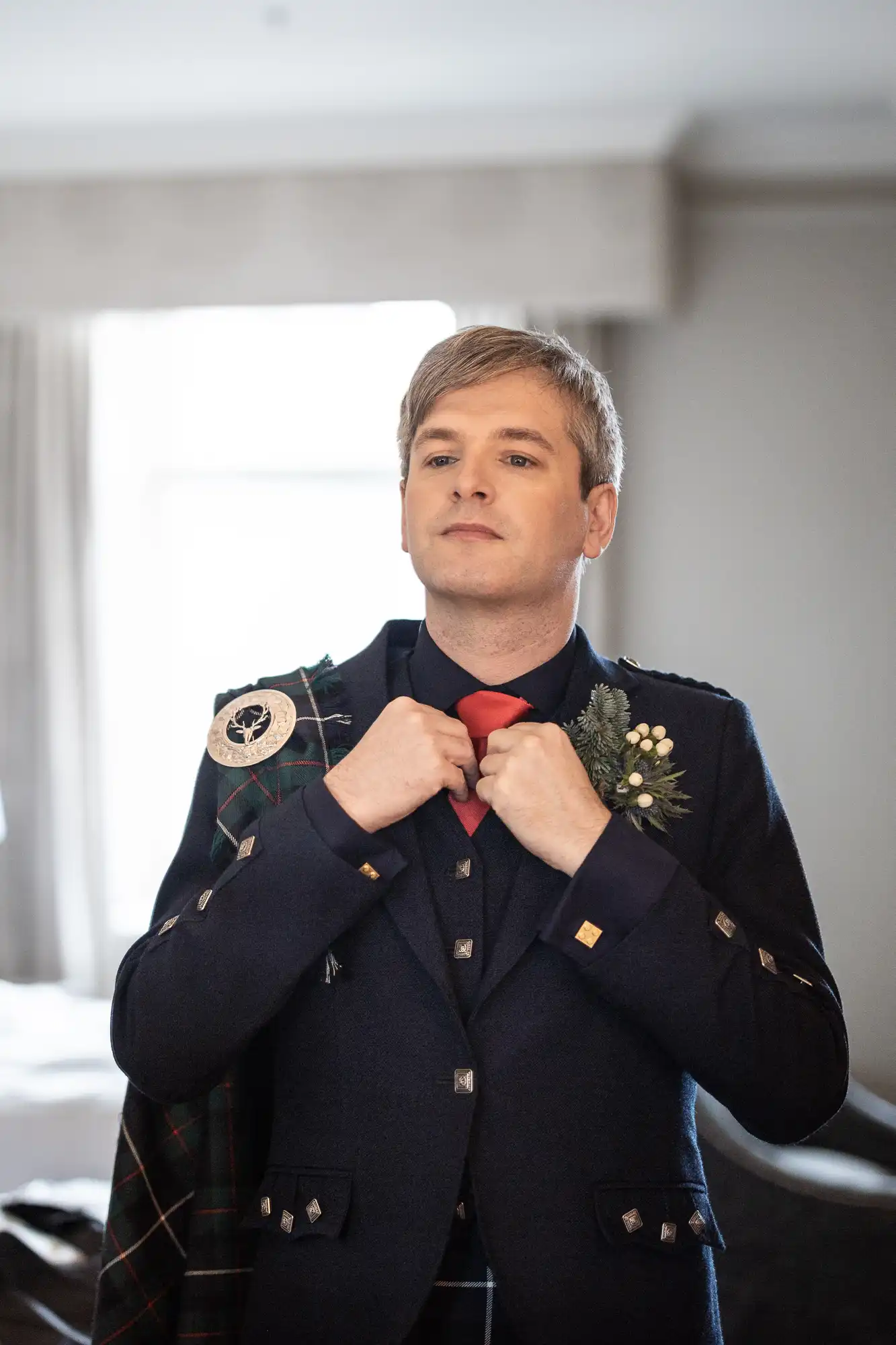A person dressed in traditional Scottish attire adjusts their red tie while standing in a room with light-colored curtains and a window in the background.