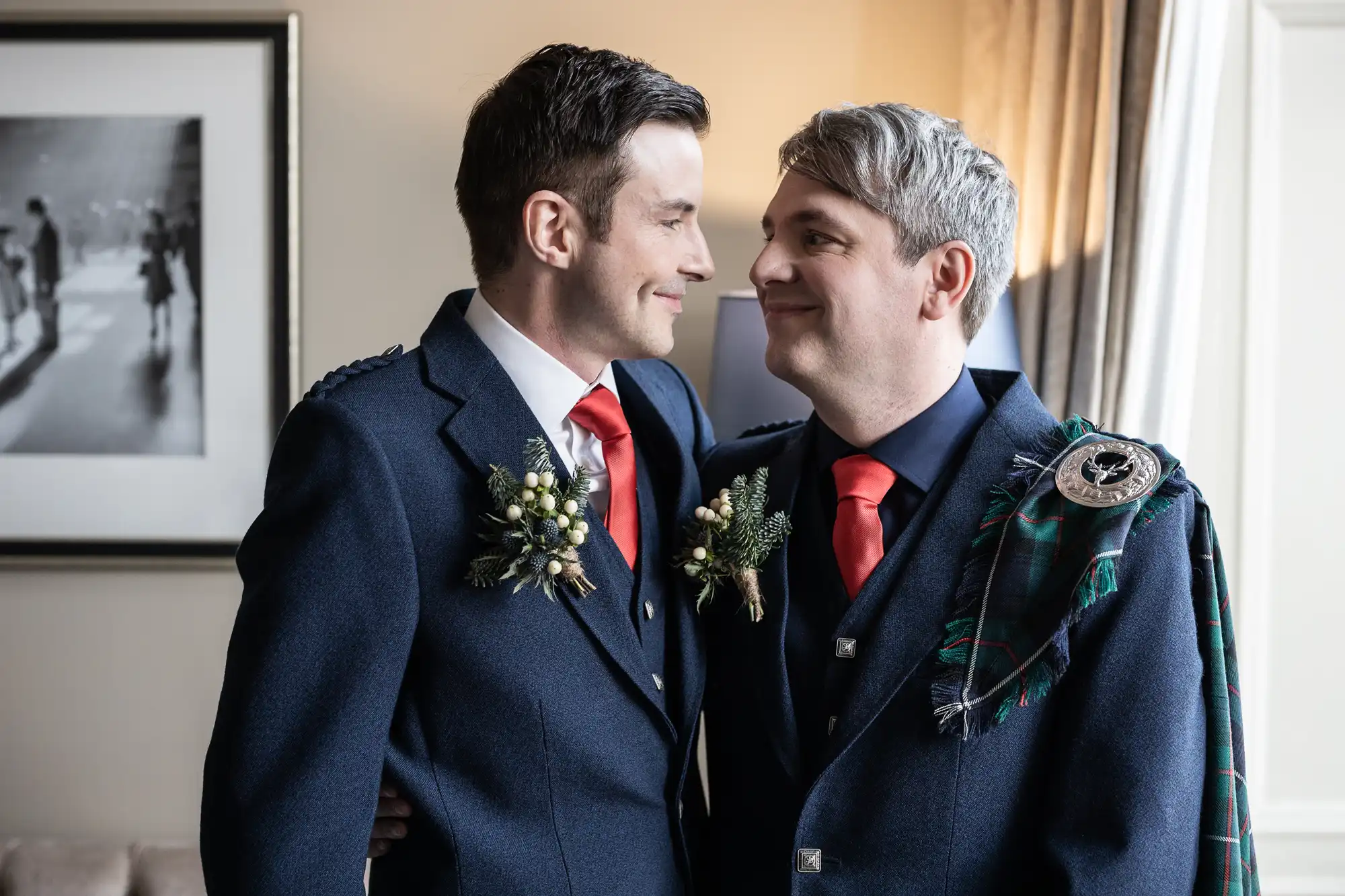 Two men, dressed in matching formal attire with red ties and floral boutonnieres, smile at each other while standing in an indoor setting with a framed photo on the wall in the background.