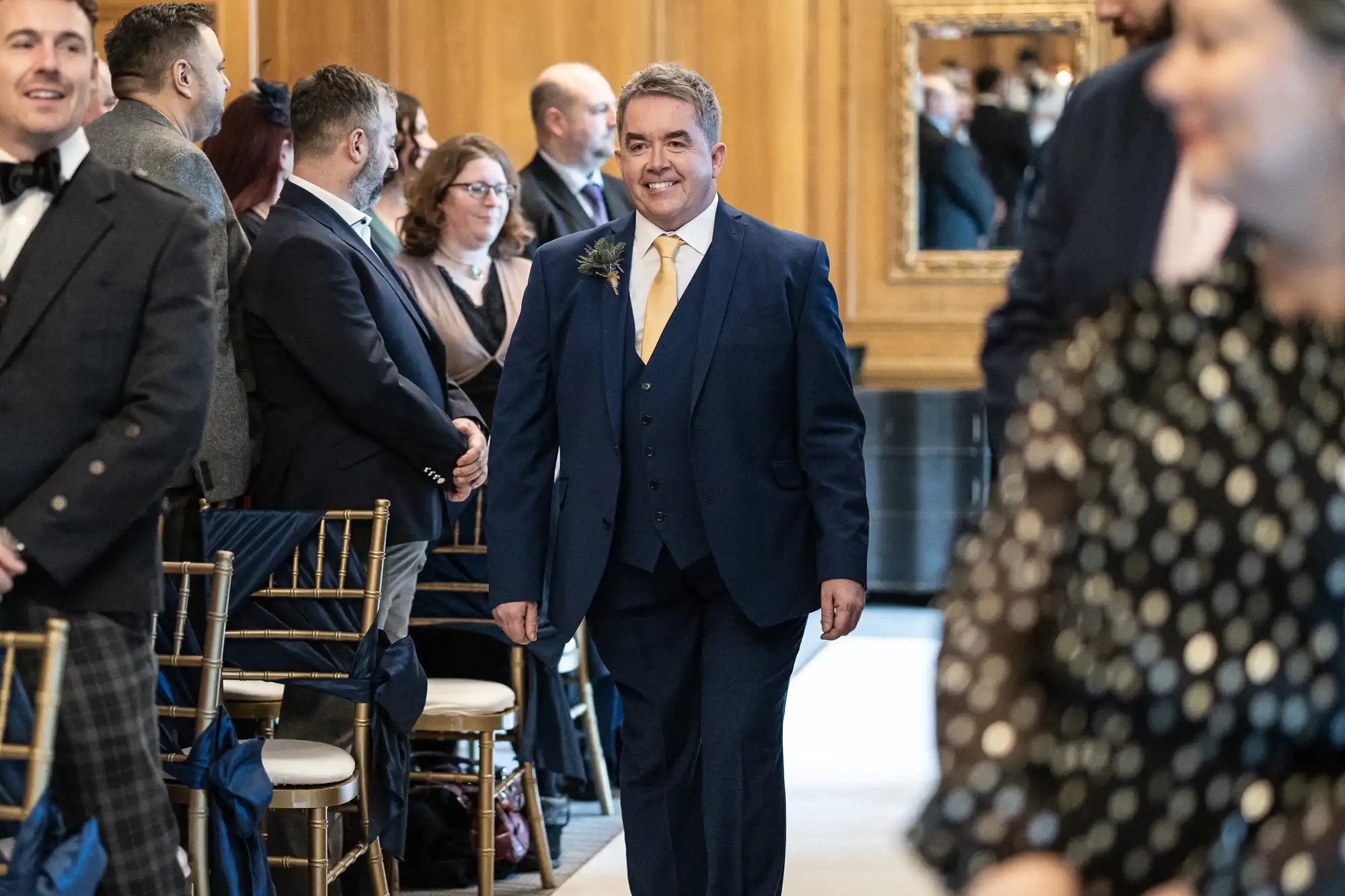 A man wearing a blue suit and yellow tie walks down an aisle in a formal setting, surrounded by seated and standing guests.