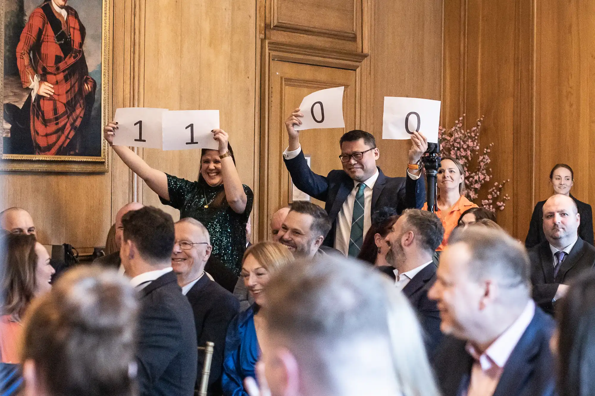 Two individuals hold up signs displaying the numbers "1100" while an audience watches, seated in a room with wooden paneling and a wall portrait.