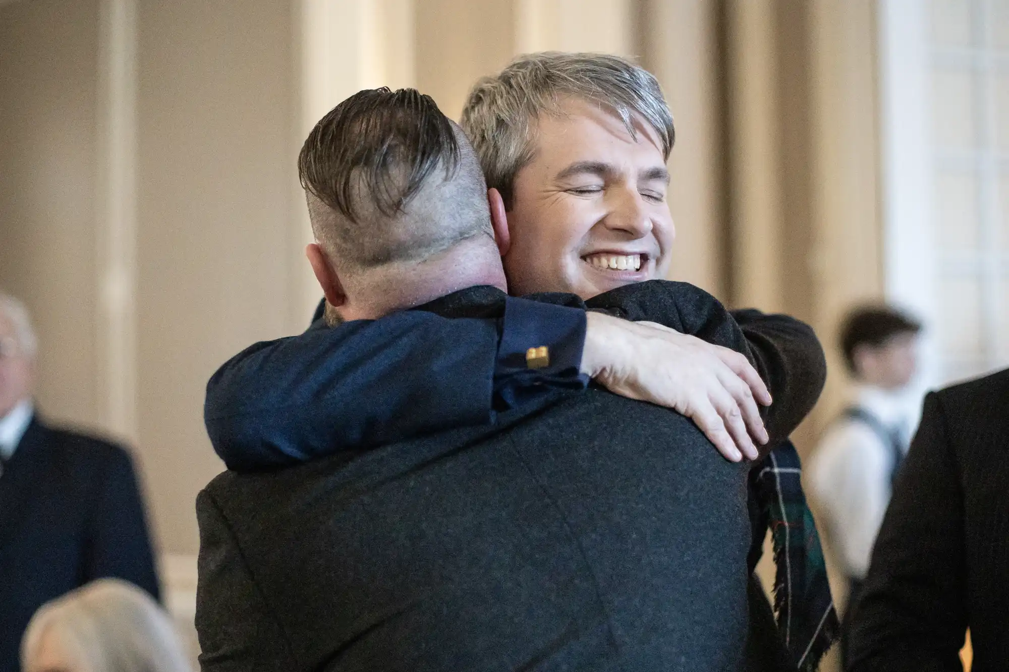 Two people are embracing warmly indoors. One person's face is visible, showing a smile, while the other person's back is turned to the camera.