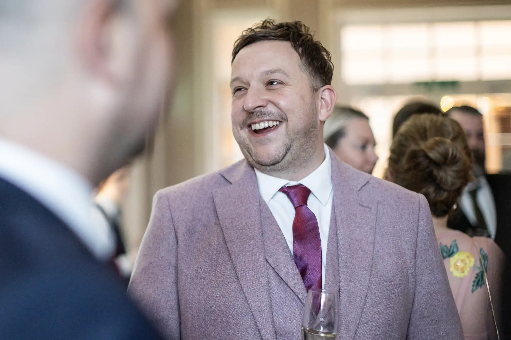 A man in a pink suit and red tie, holding a glass, smiles and talks with others at a social event. People in formal attire are visible in the background.