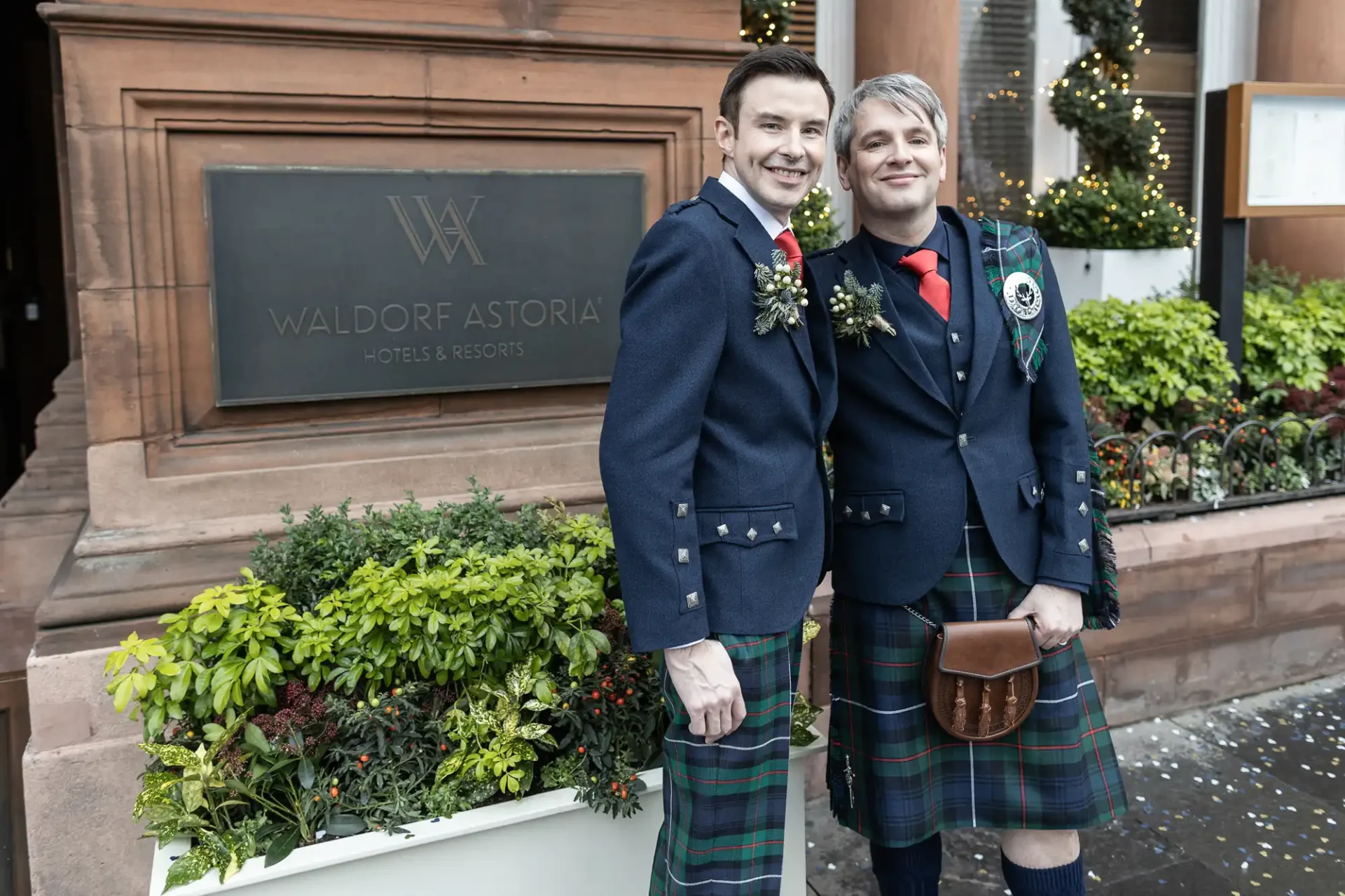 The Caledonian Edinburgh weddings image of Two men wearing traditional Scottish kilts stand side by side smiling outside the Waldorf Astoria hotel, with festive decorations in the background.