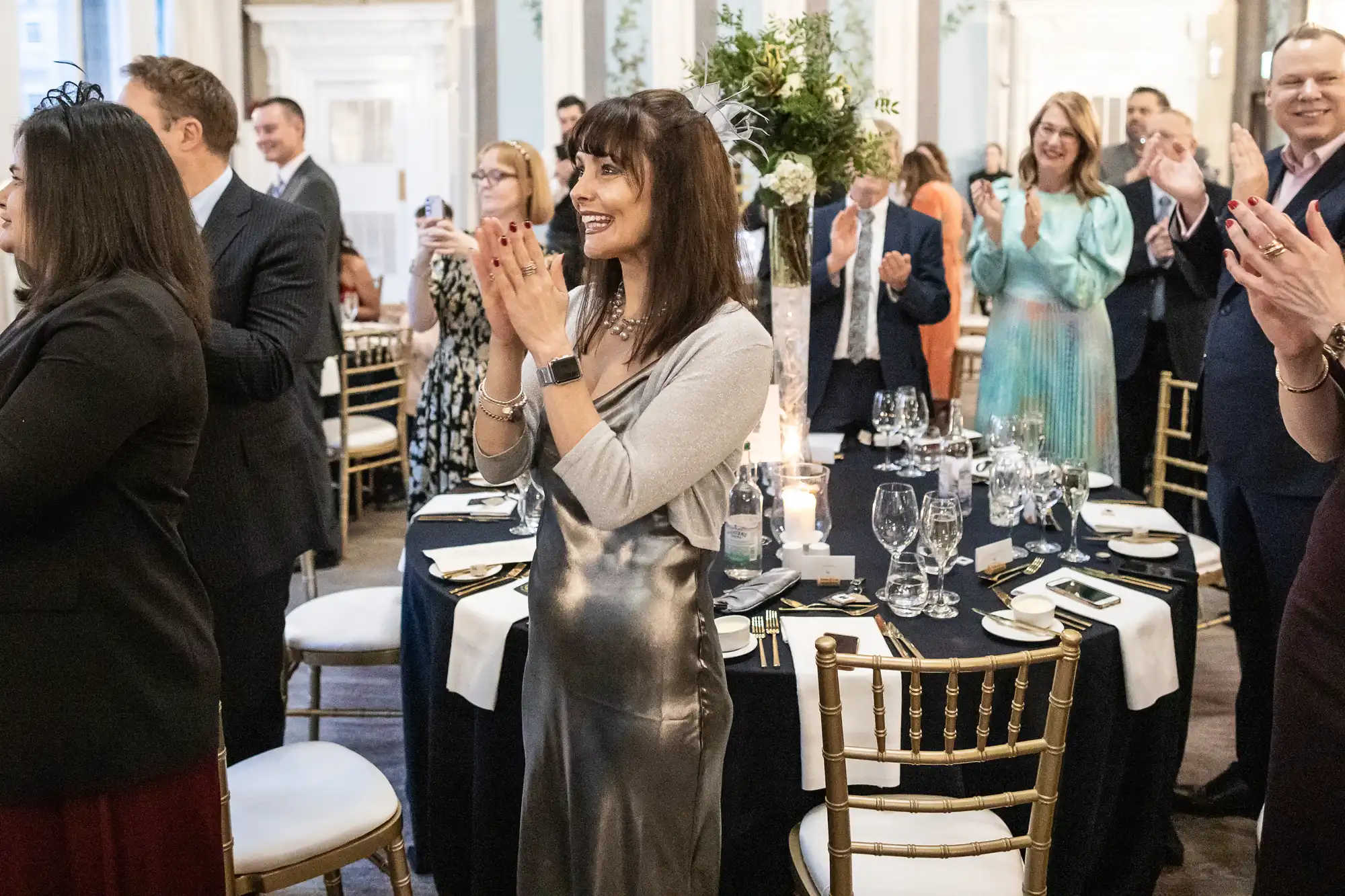 People standing and clapping at a formal event in a decorative room with round tables set with glassware and candles.
