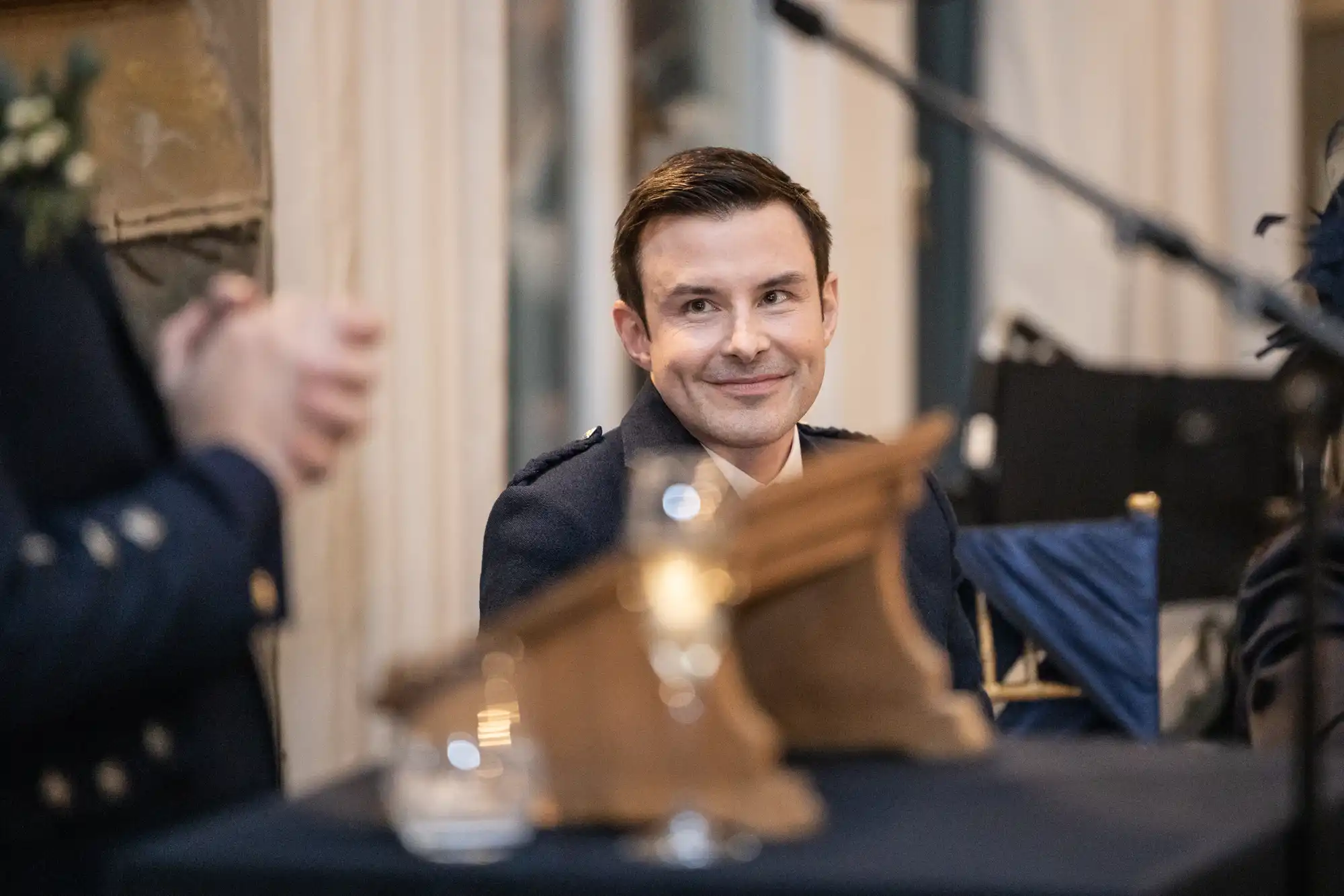A man in a suit sits smiling, listening attentively near a wooden podium with a glass of water on it. Microphones are set up nearby.